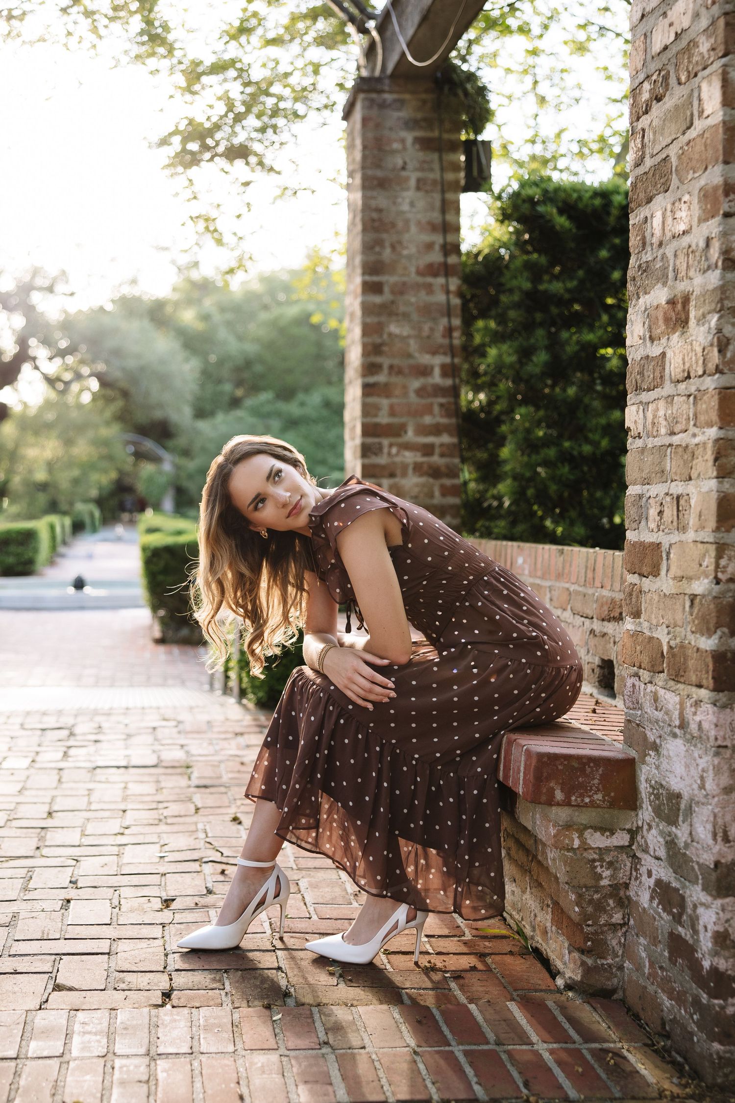 Woman in brown polka dot dress and white heels sitting on brick steps in a lush garden setting.