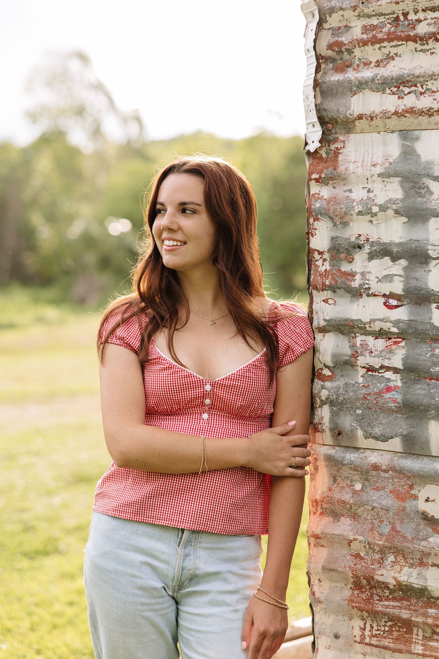 Young woman in red gingham top and jeans leaning against rustic metal wall outdoors, smiling.