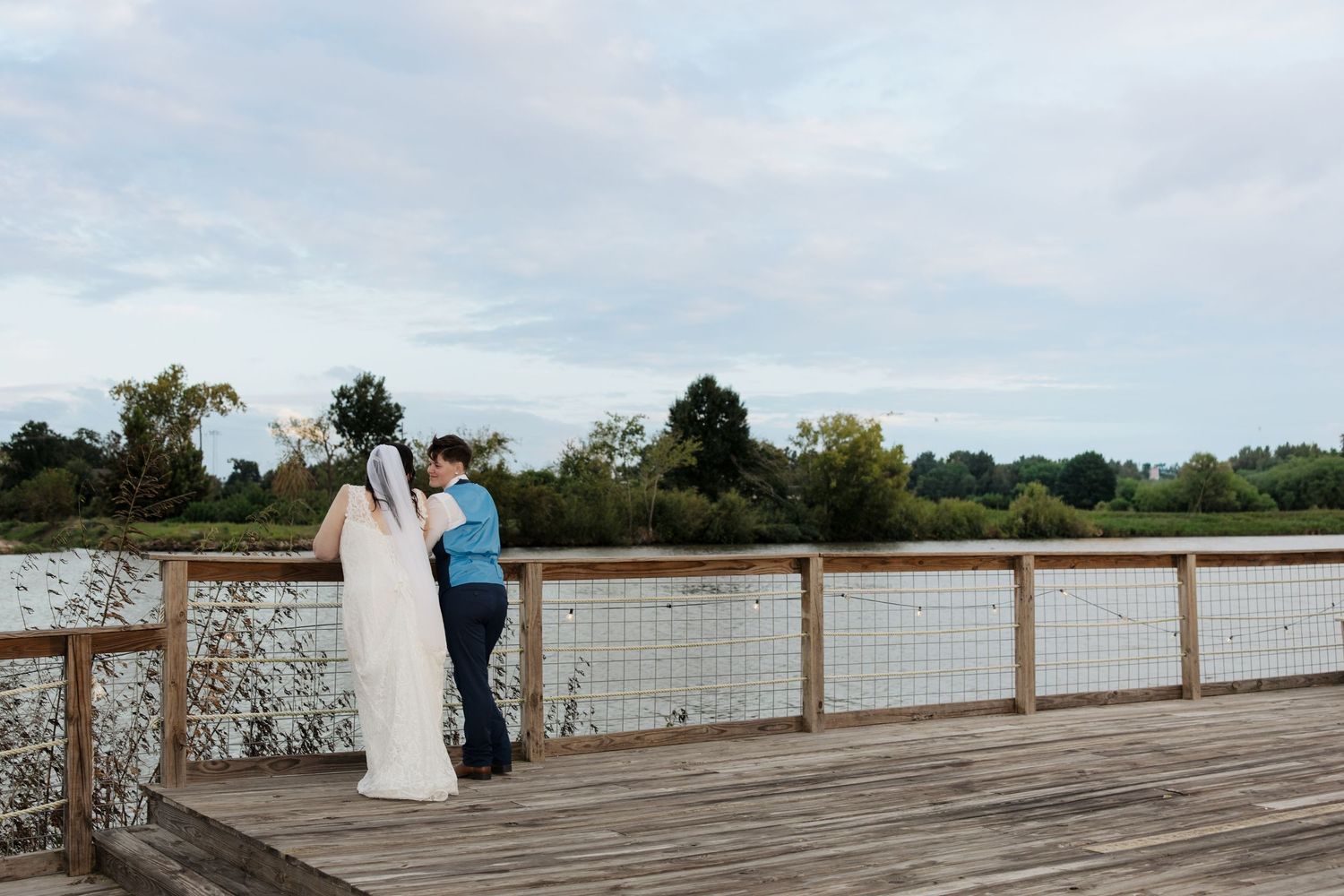 Wedding group stands on wooden deck overlooking scenic waterfront view on cloudy day.