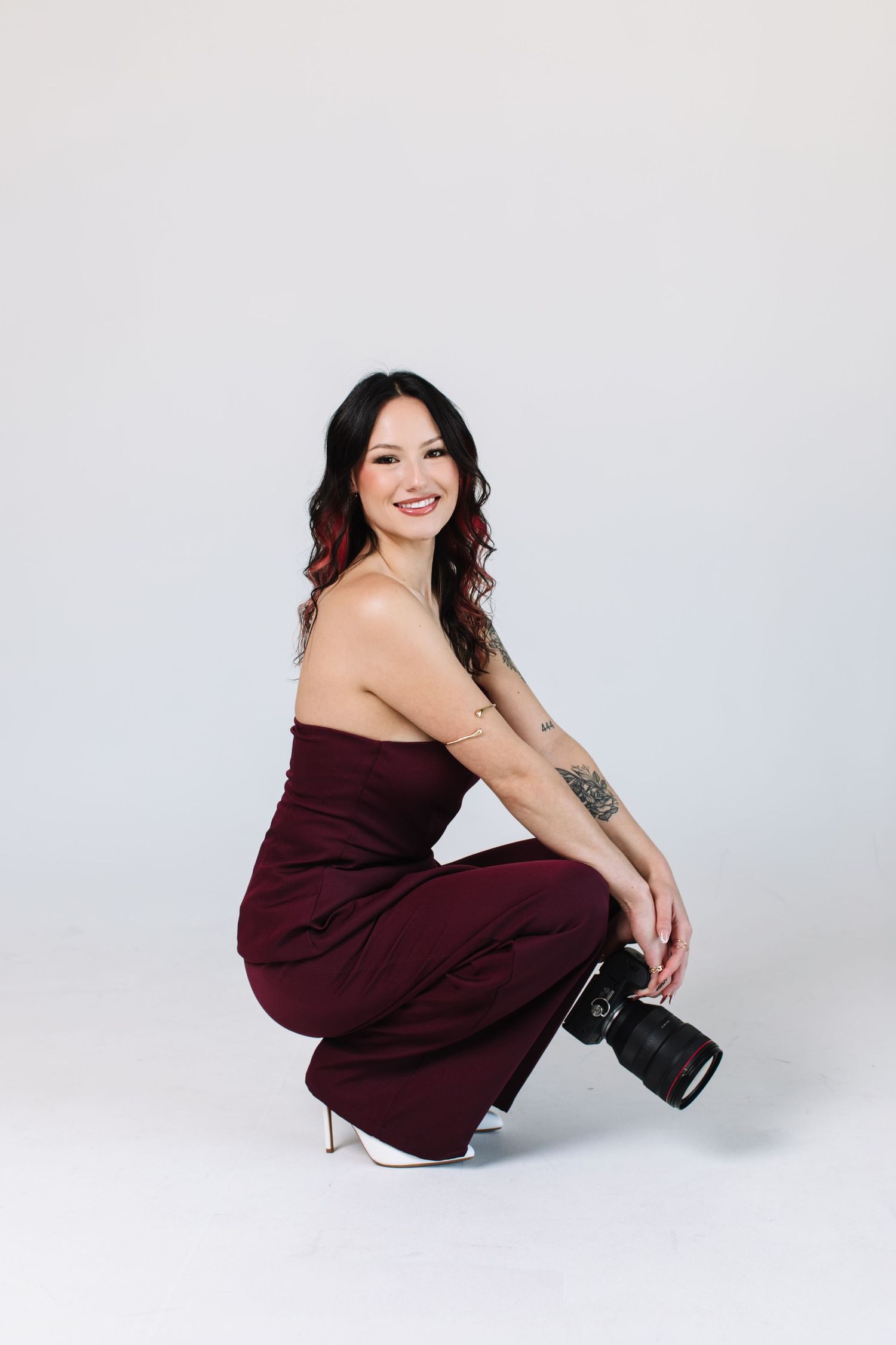 Woman in burgundy strapless jumpsuit crouching with a camera, smiling, tattoo on arm, white heels.