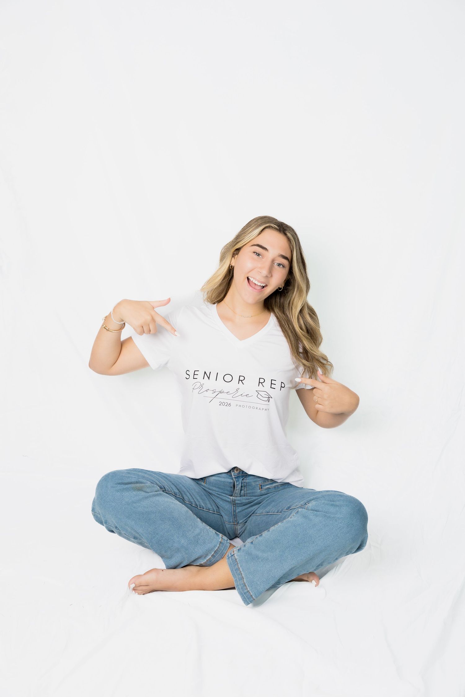Woman in jeans sits cross-legged on white background pointing to text on her t-shirt.