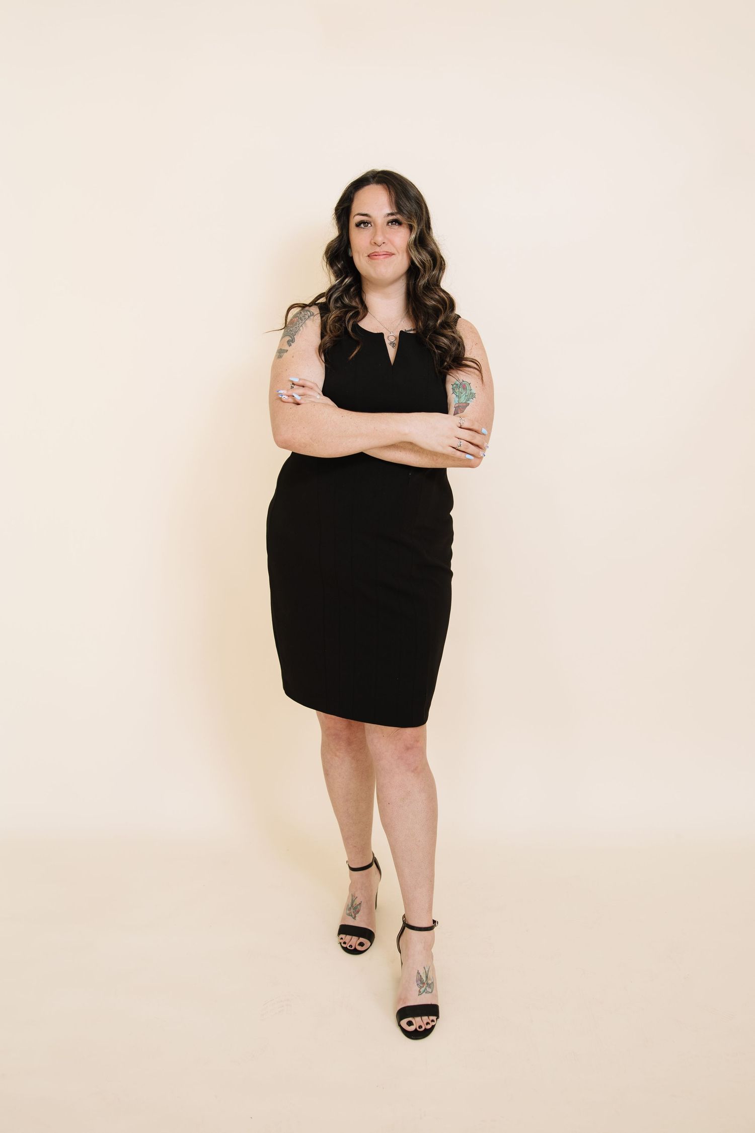 A person in a little black dress and strappy sandals poses confidently against a light colored wall.