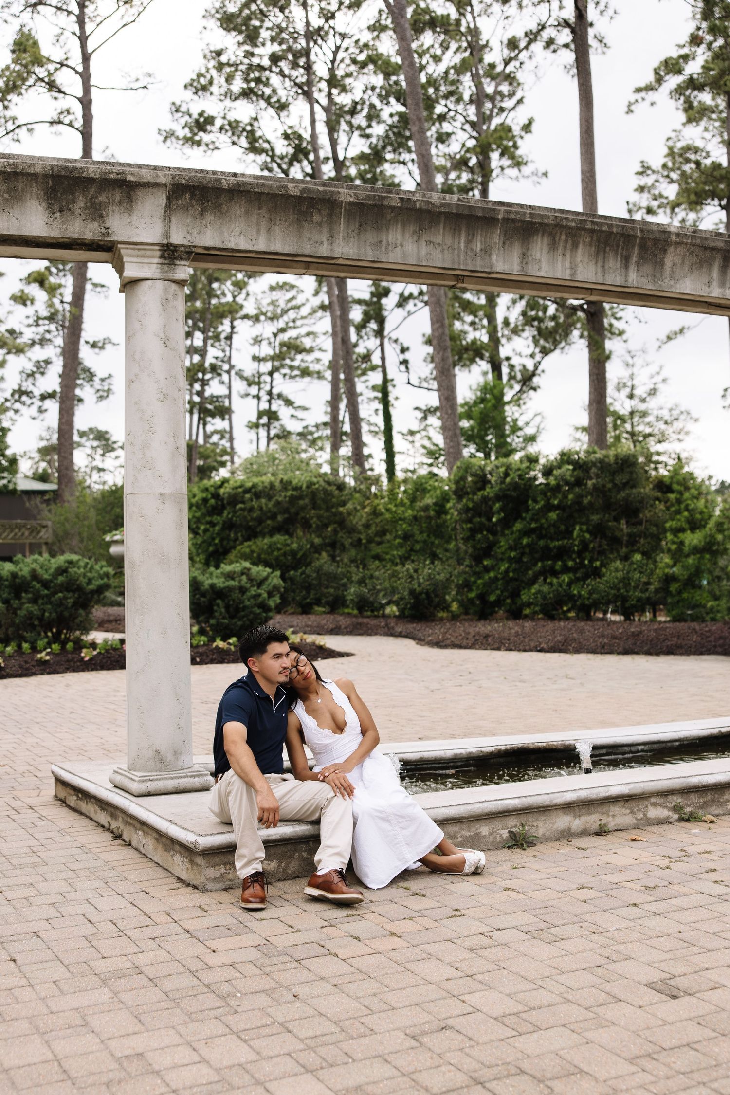 Couple sitting together on a stone bench beside a decorative column in a lush garden park setting.