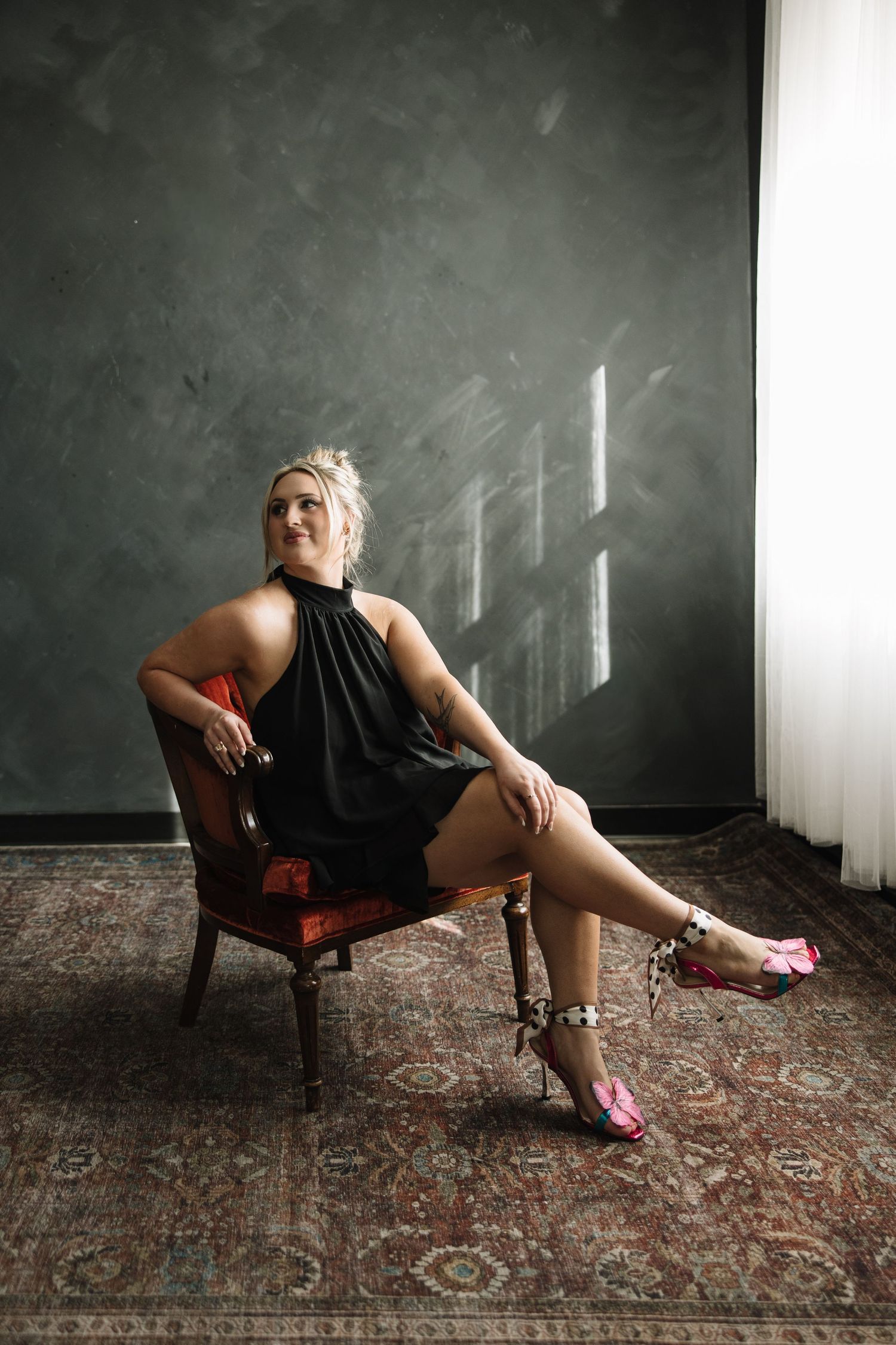 Figure in black dress and pink shoes reclining on vintage chair in moody room with dramatic lighting.