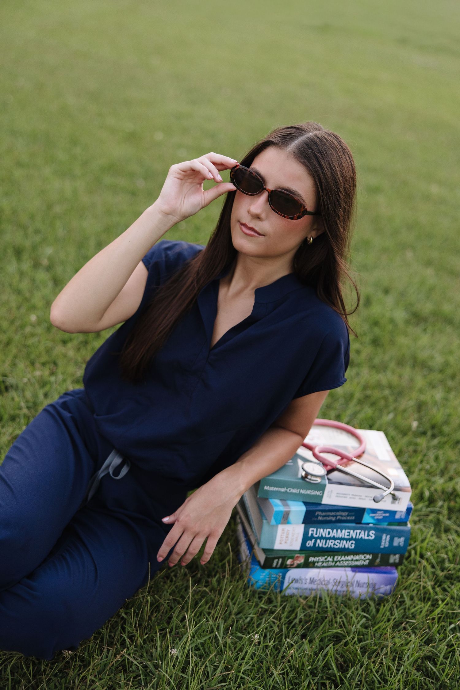 Nursing student in navy scrubs sitting on grass with Fundamentals of Nursing textbooks and stethoscope, adjusting sunglasses.