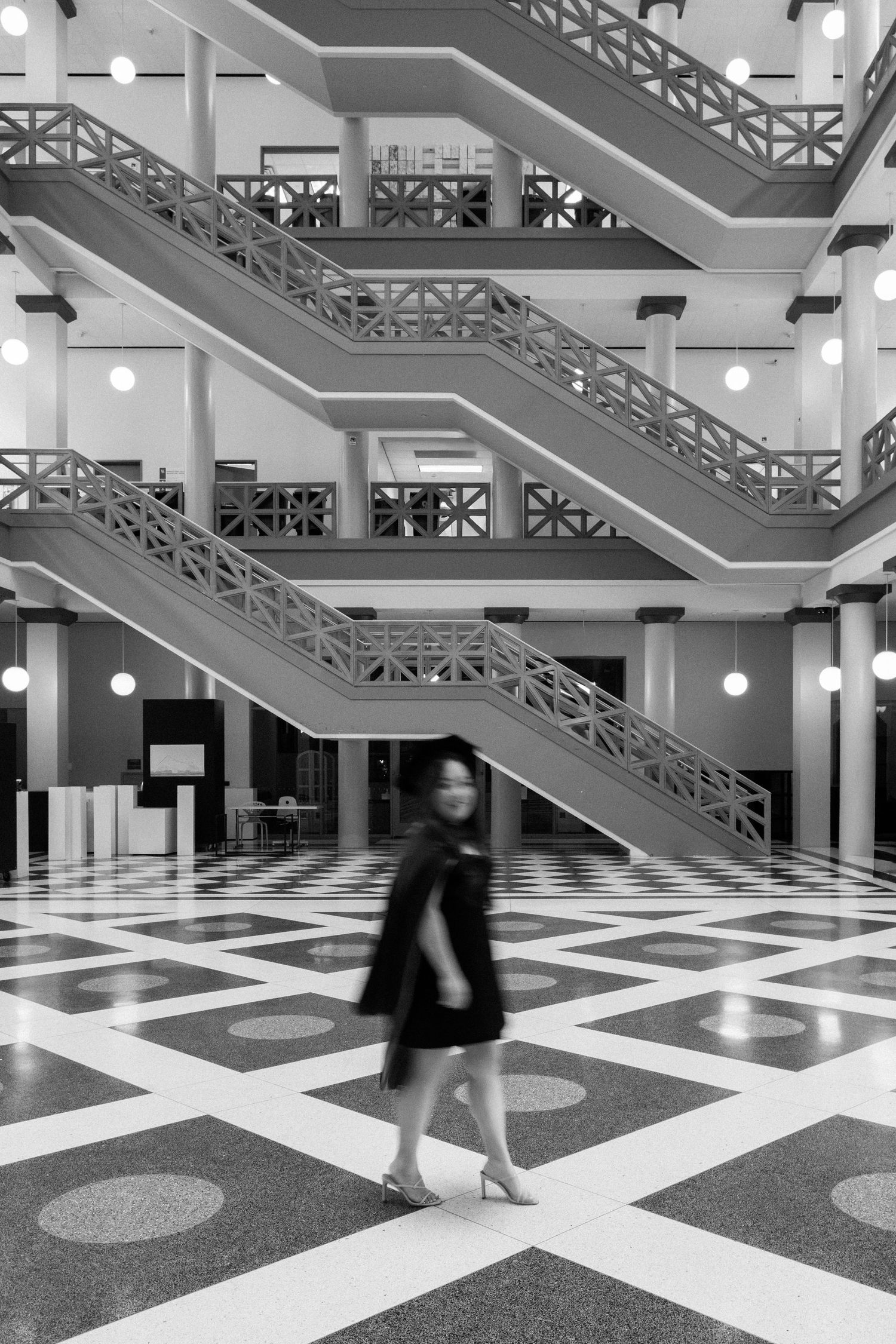 Black and white photo of a blurred woman walking across a checkered floor in a grand multi-level building with staircases.