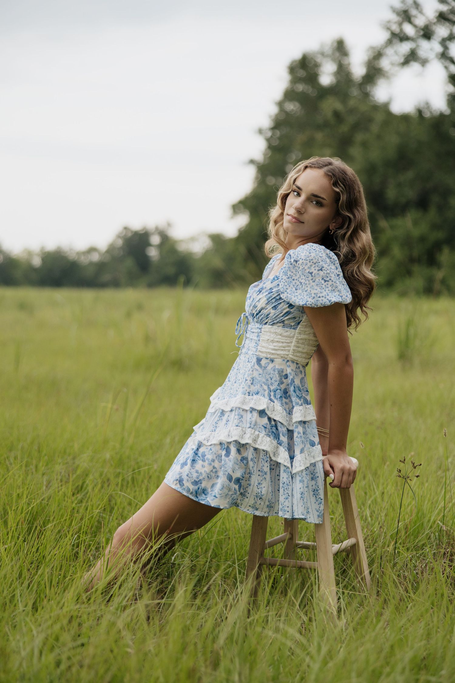 Young woman in blue floral dress poses on wooden stool in summer meadow.