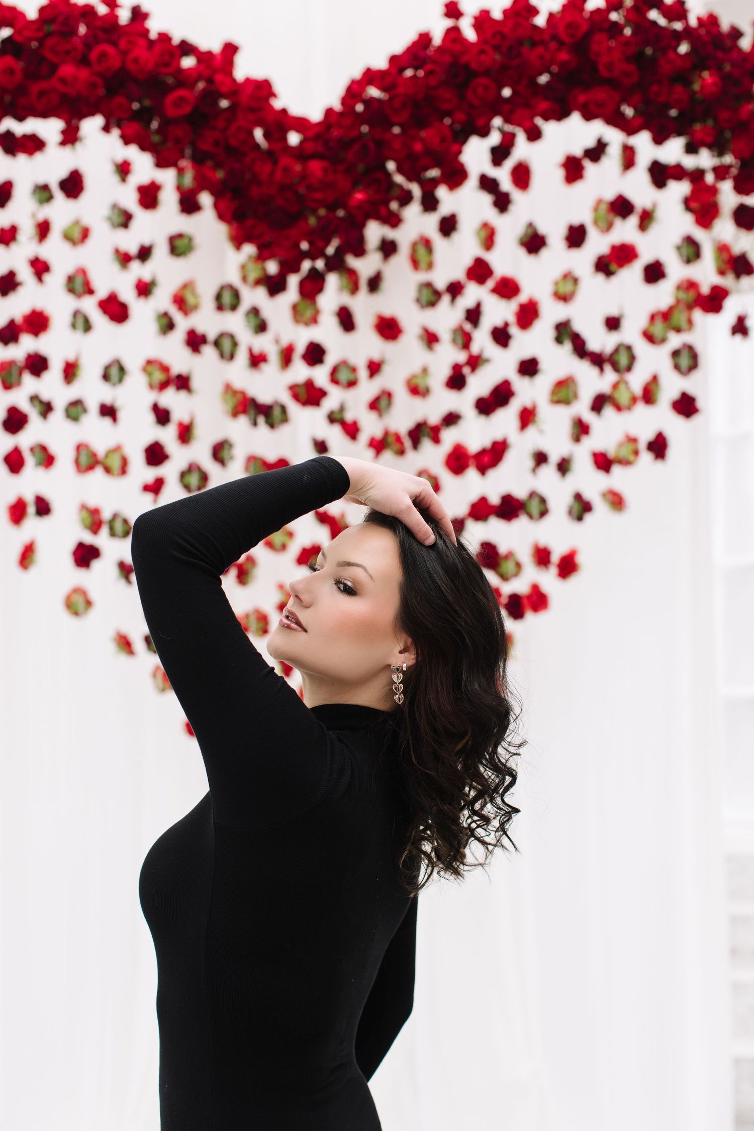 Woman in black turtleneck poses against white wall with cascading red rose petals creating dramatic contrast.