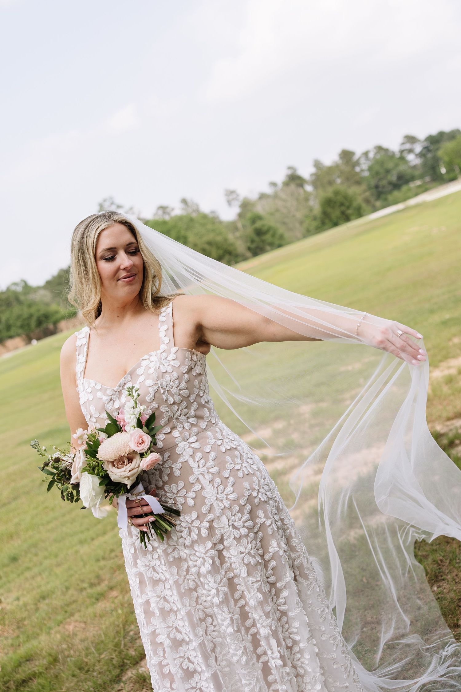 Bride in floral lace gown holding pink bouquet, veil flowing in breeze on green outdoor wedding venue.