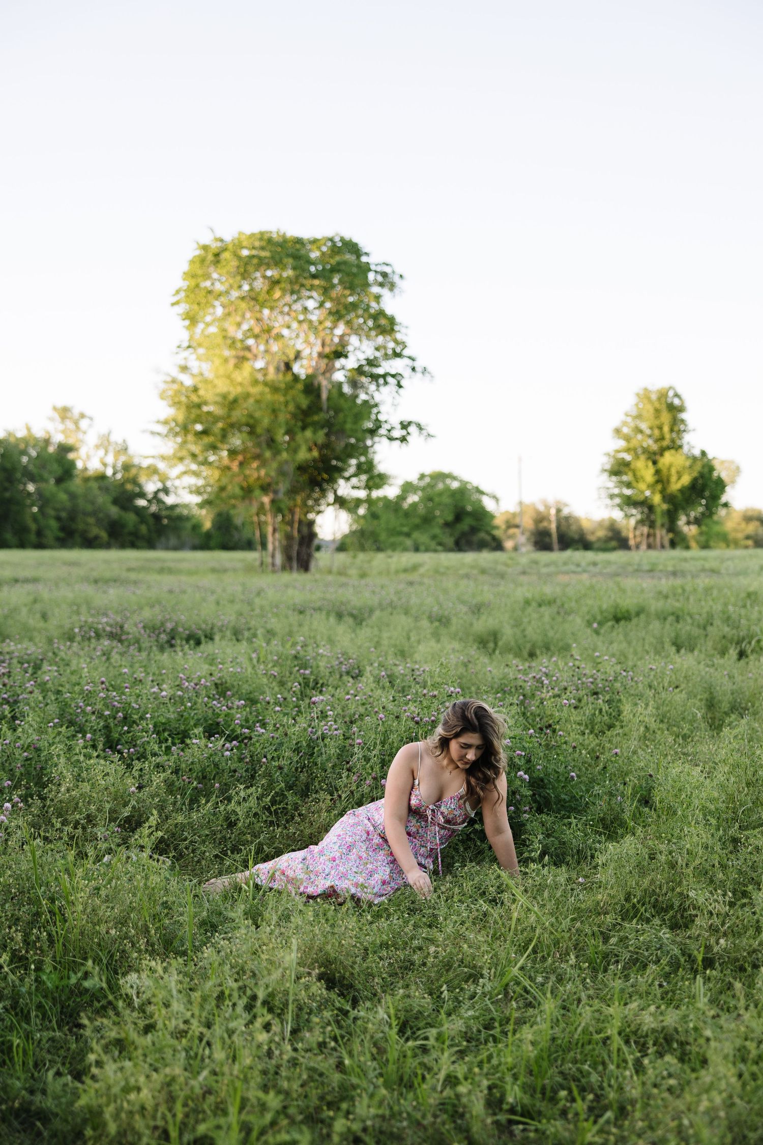 Woman in floral dress sitting in lush green field at golden hour with trees in background.