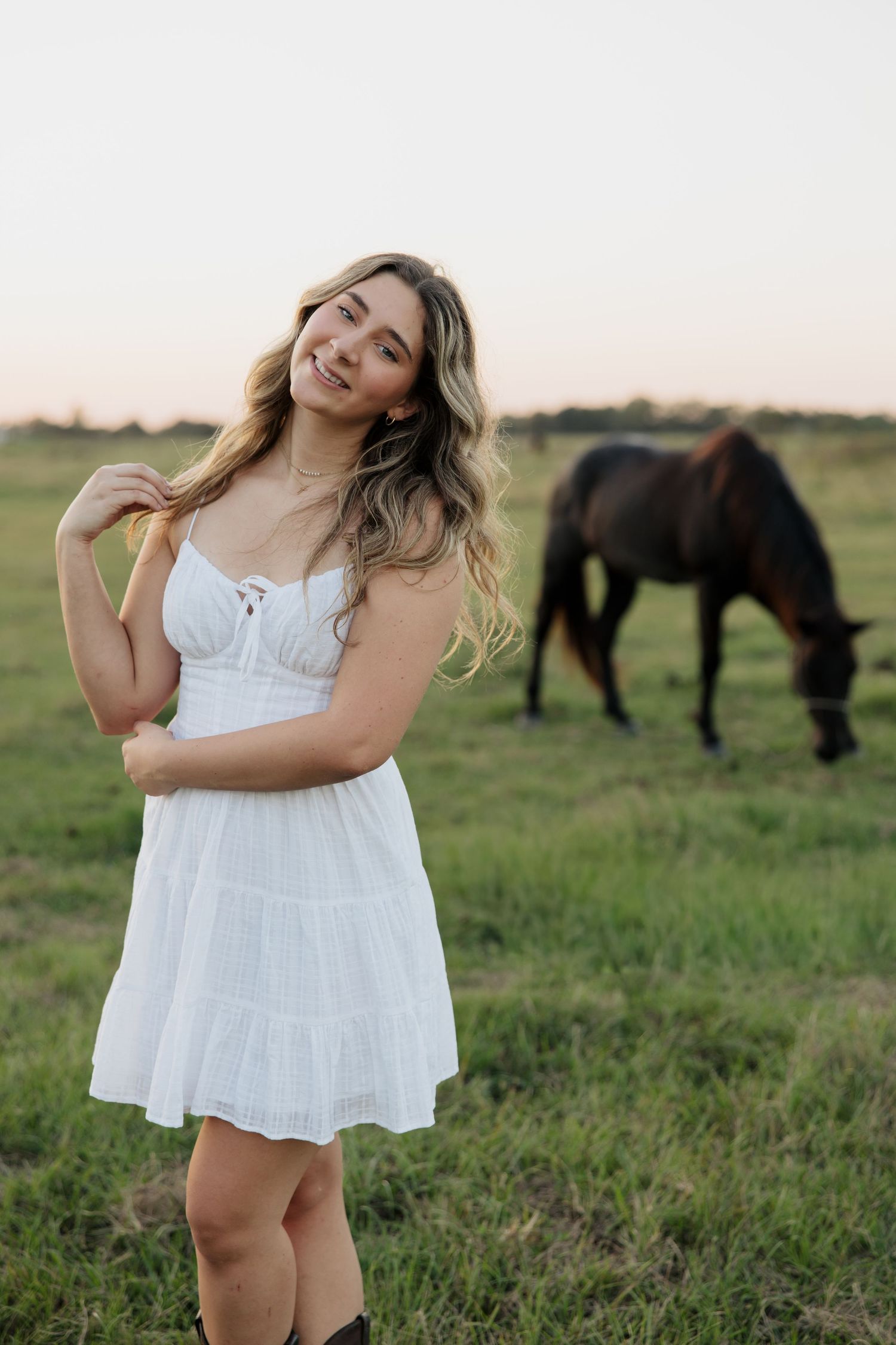 Young woman in white sundress smiling in a green field with a brown horse grazing in the background.