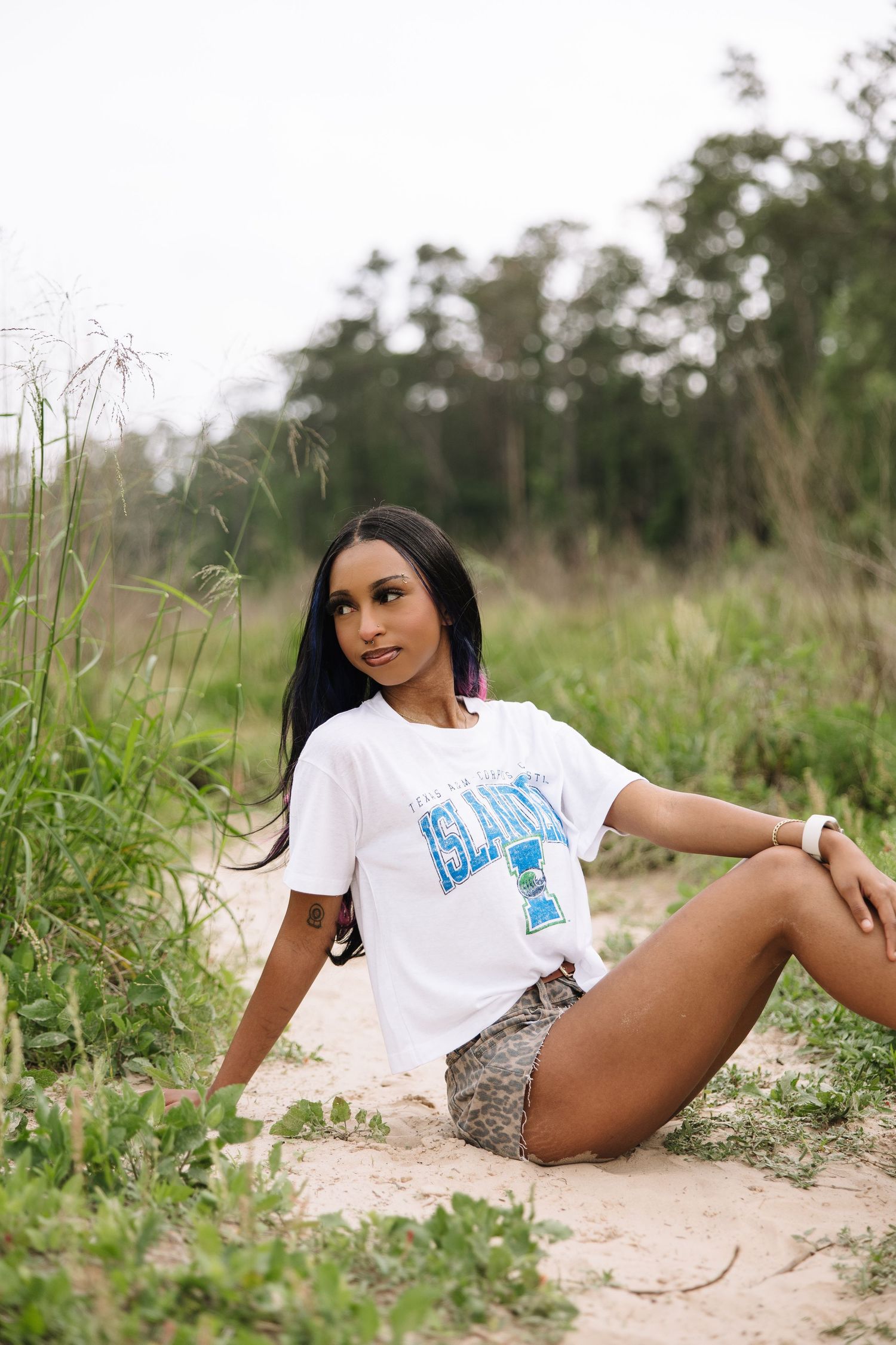 Young woman in white graphic tee and camo shorts sitting on sandy path surrounded by lush green vegetation outdoors.