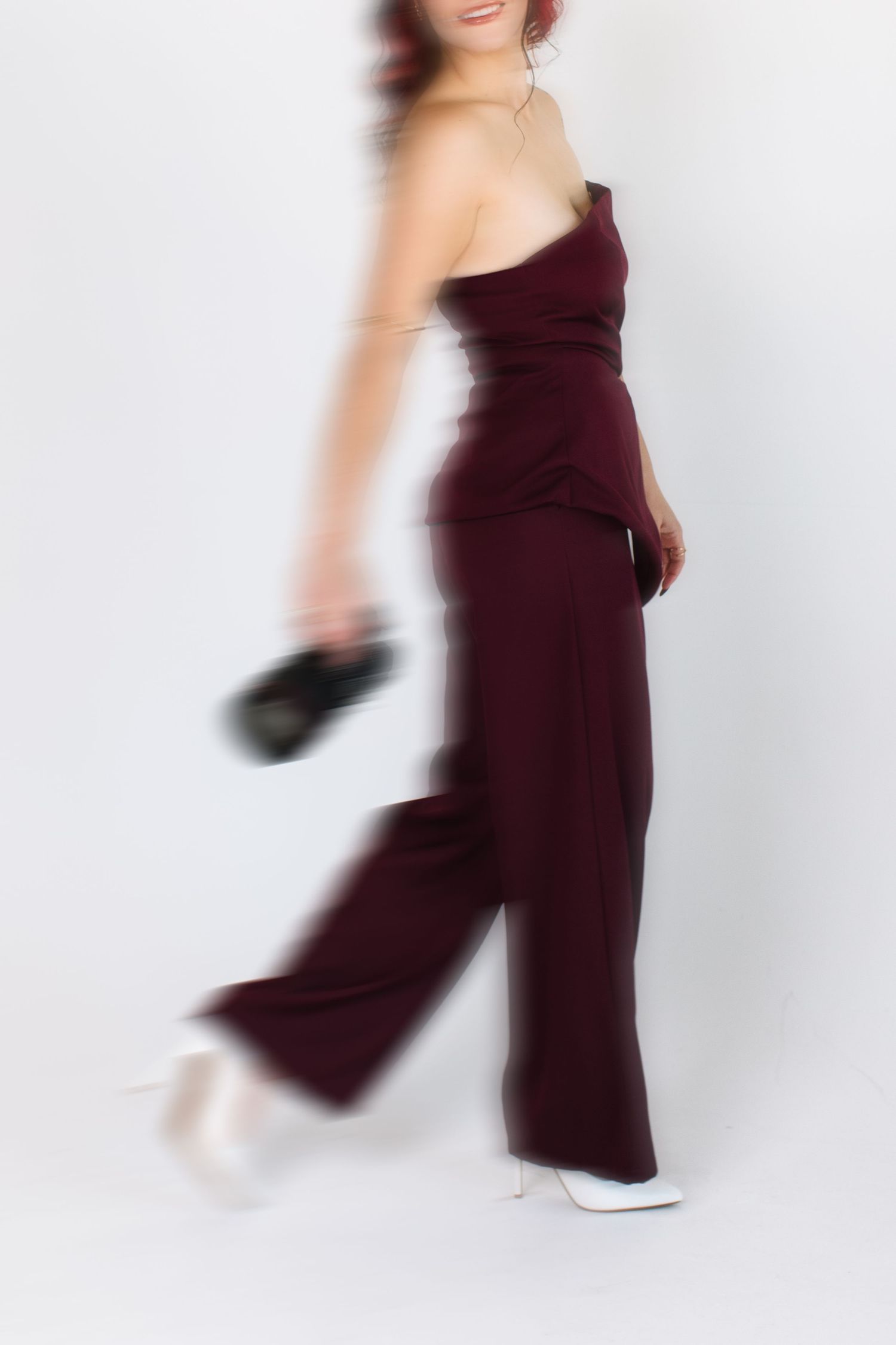 Woman in burgundy one-shoulder jumpsuit and white heels holding a black clutch, motion blur effect.