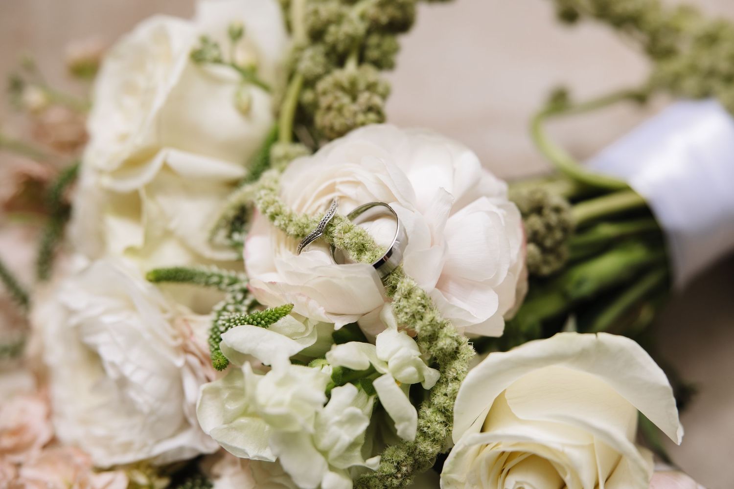 Wedding rings nestled among white ranunculus and roses in an elegant bridal bouquet with green accents.