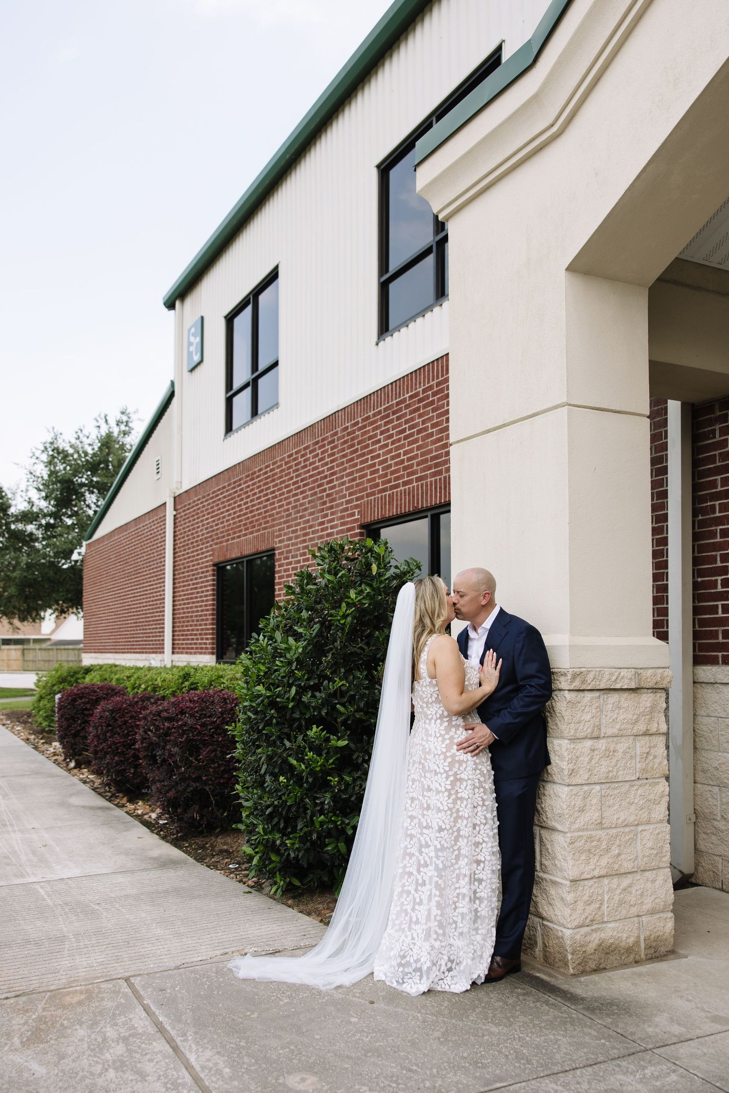 Bride and groom share a romantic kiss against a brick pillar outside a building, with her lace gown and veil trailing.