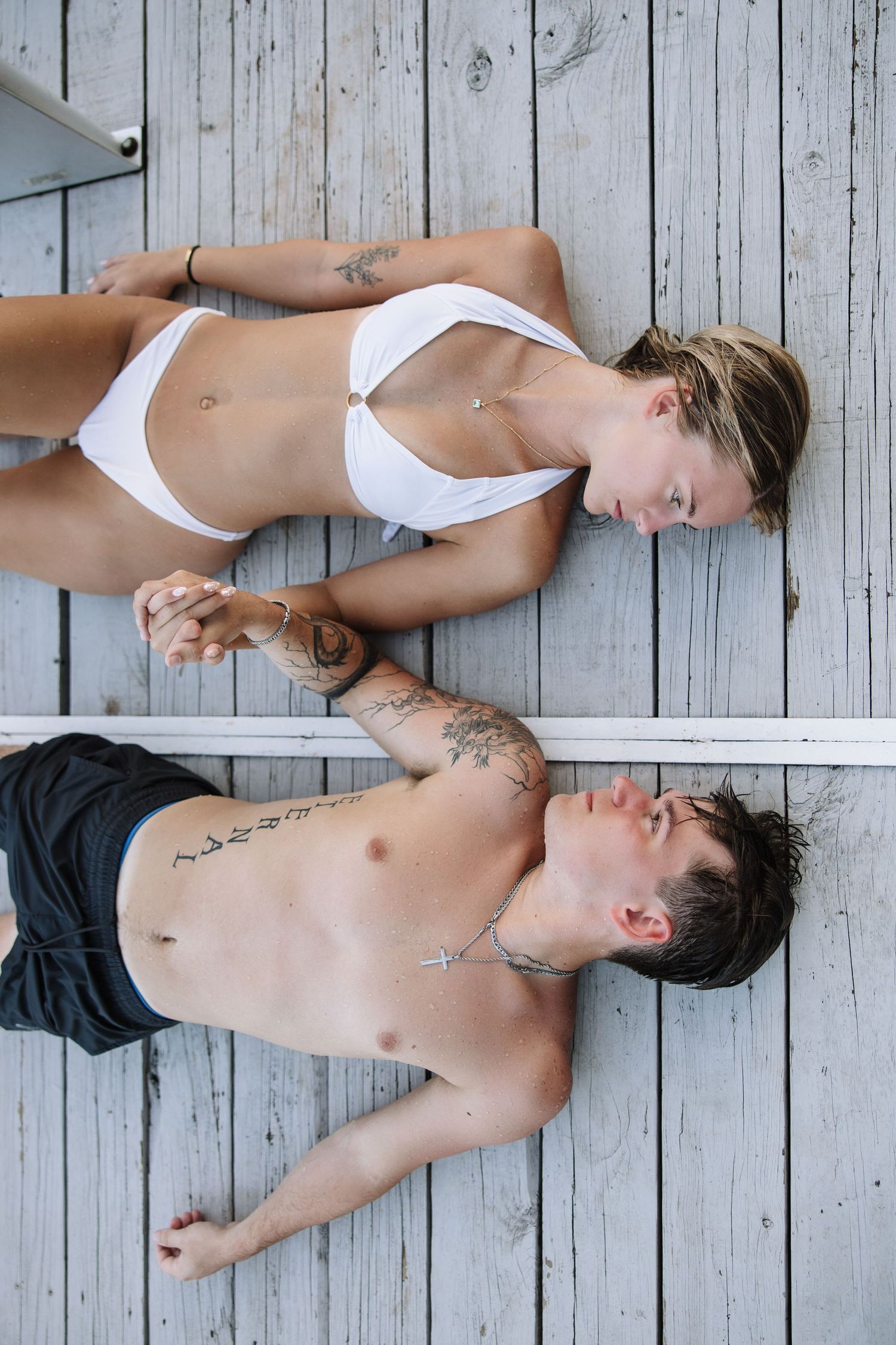 Two tattooed young adults in swimwear lying on a wooden dock, holding hands and relaxing together outdoors.
