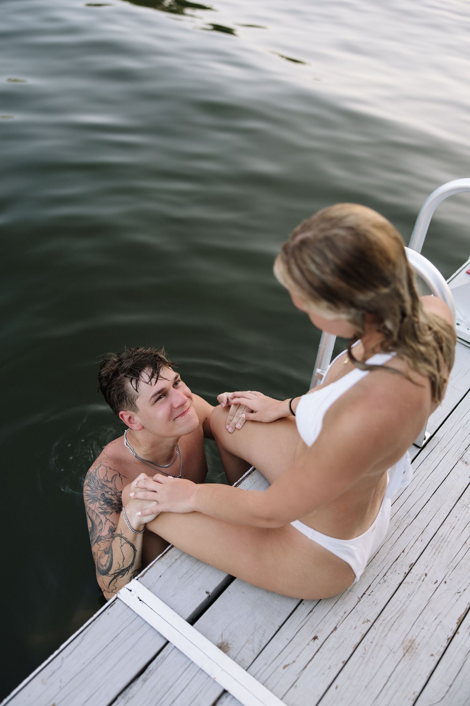 Two people sharing a romantic moment on a wooden dock, one in the water with tattoos, the other in a white bikini.