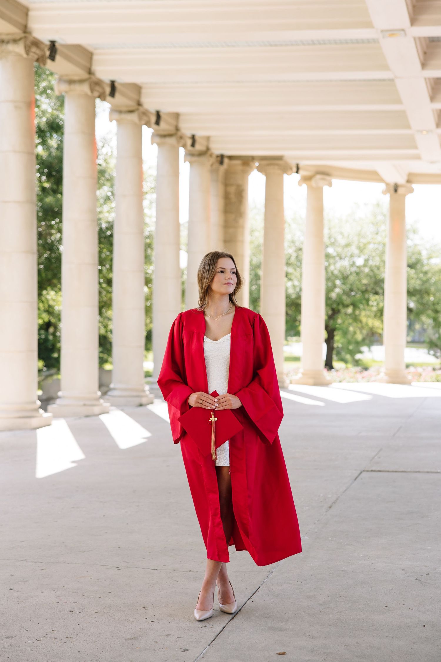 Young woman in red graduation gown poses between classical columns outdoors, holding tassel in natural light.