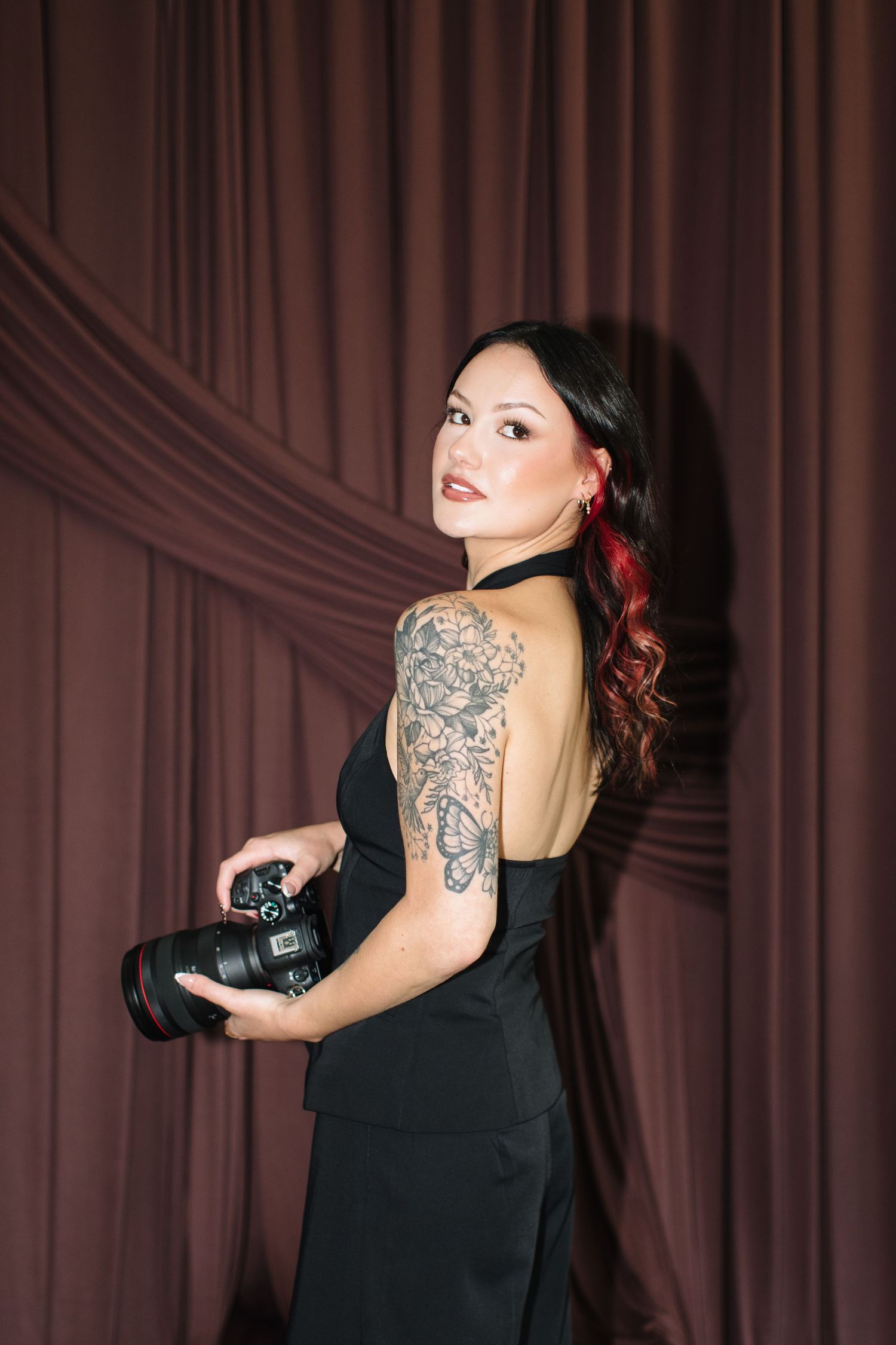 Woman in black backless dress with floral arm tattoo holding a camera, posing against burgundy drape backdrop.