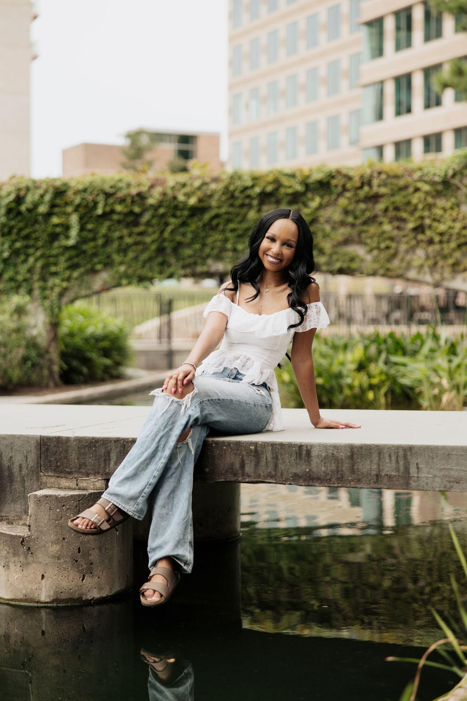 Woman in white top and jeans relaxes by a reflective pond in an urban garden setting.