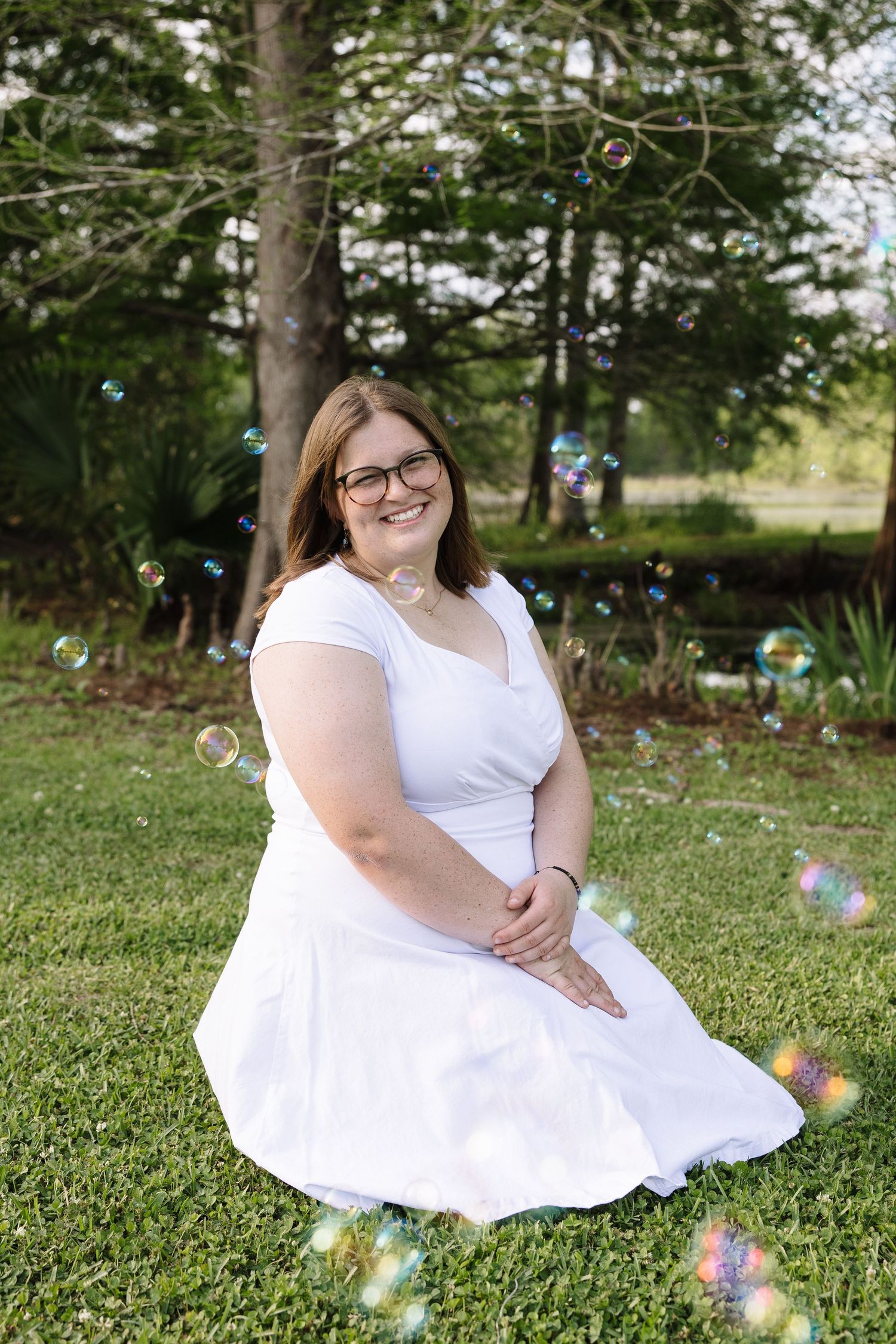 Young woman in white dress sitting on green grass surrounded by floating bubbles in a sunny outdoor park setting.