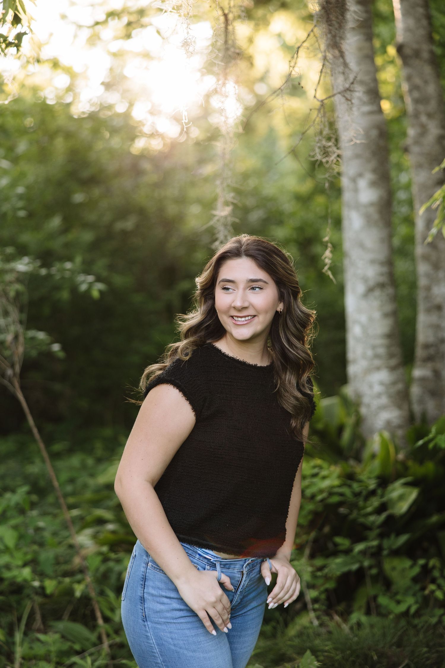 Young woman in black top and jeans smiling outdoors in lush green forest with golden sunlight filtering through trees.