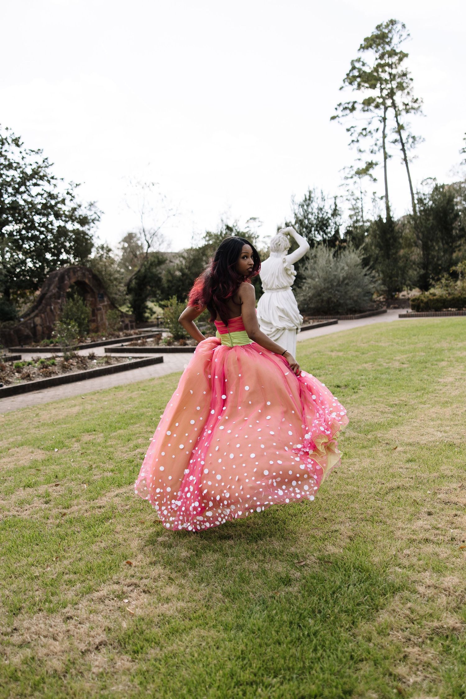 Young woman twirling in a stunning pink floral ball gown in a lush garden with a white statue backdrop.