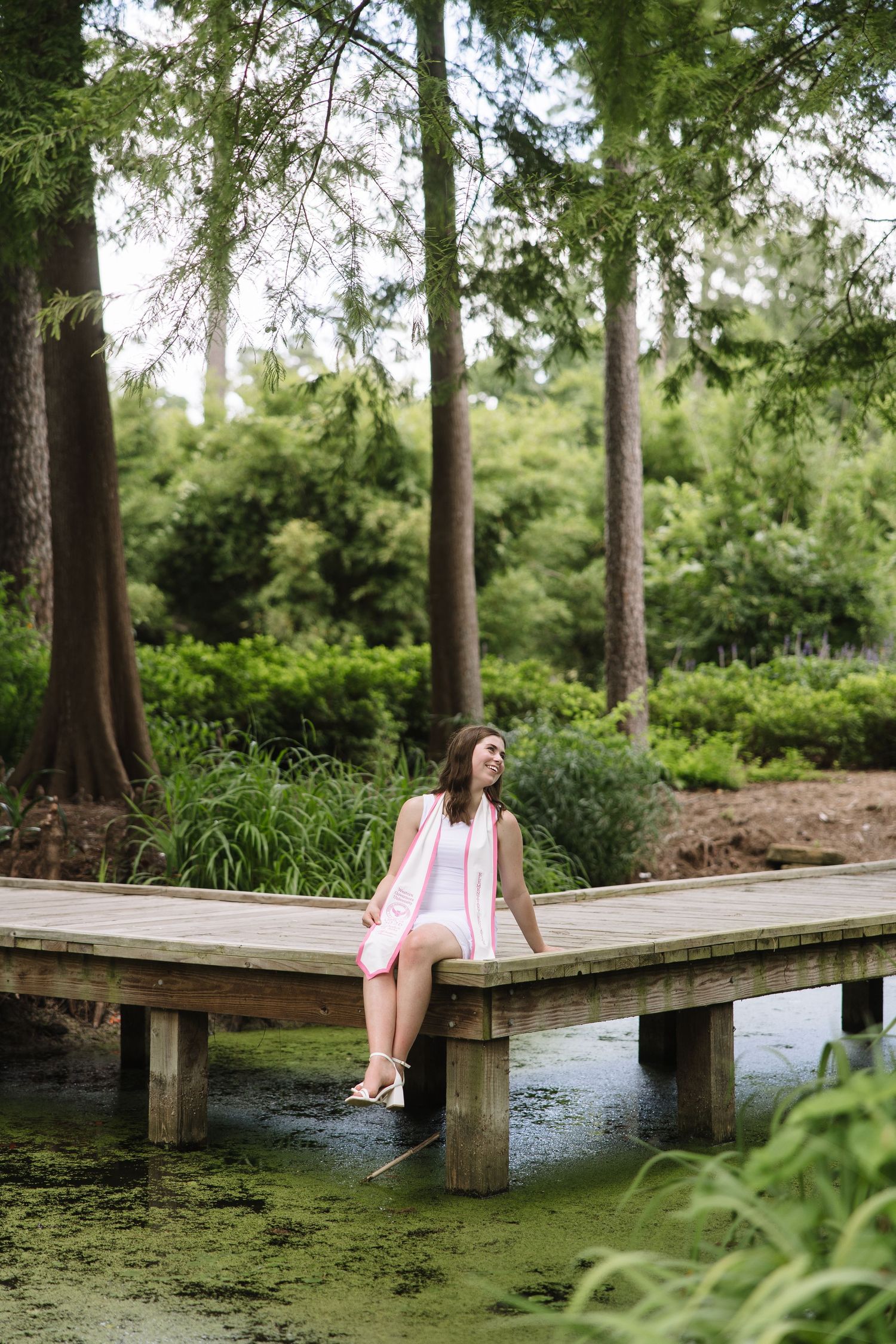 Young woman in pink dress sitting on wooden dock surrounded by lush green trees and pond.