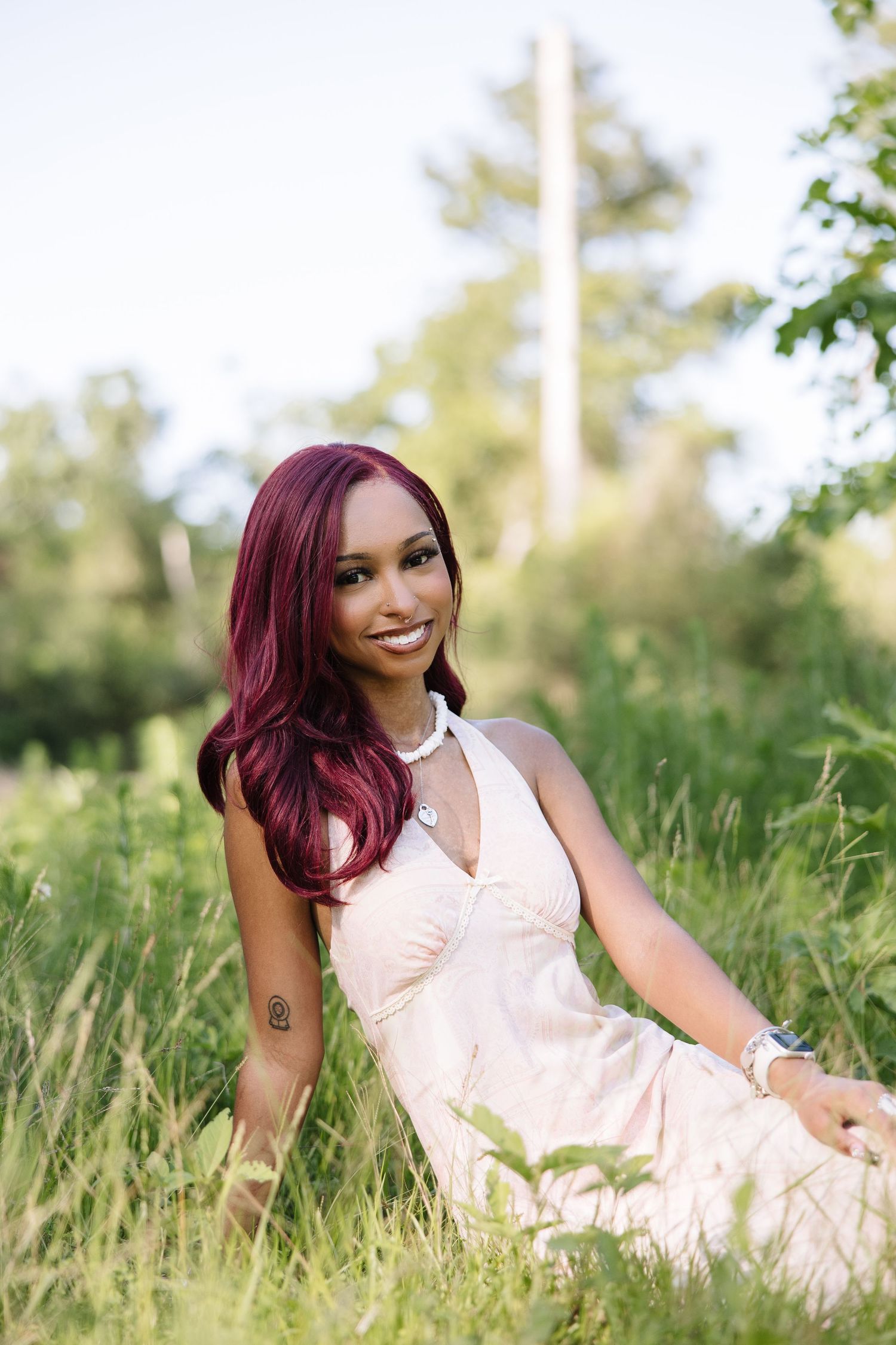 A smiling woman with red hair in a pink dress sits in lush green grass outdoors.