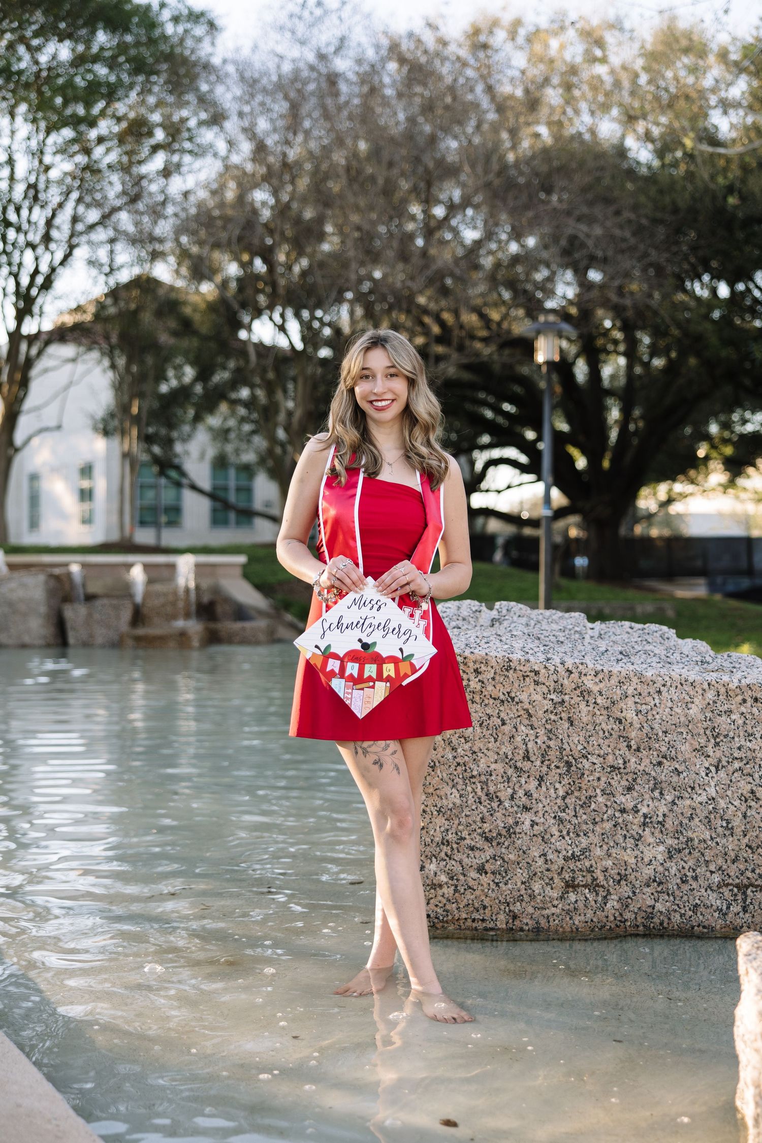 Young woman in red dress standing in fountain holding graduation cap sign, celebrating outdoors.