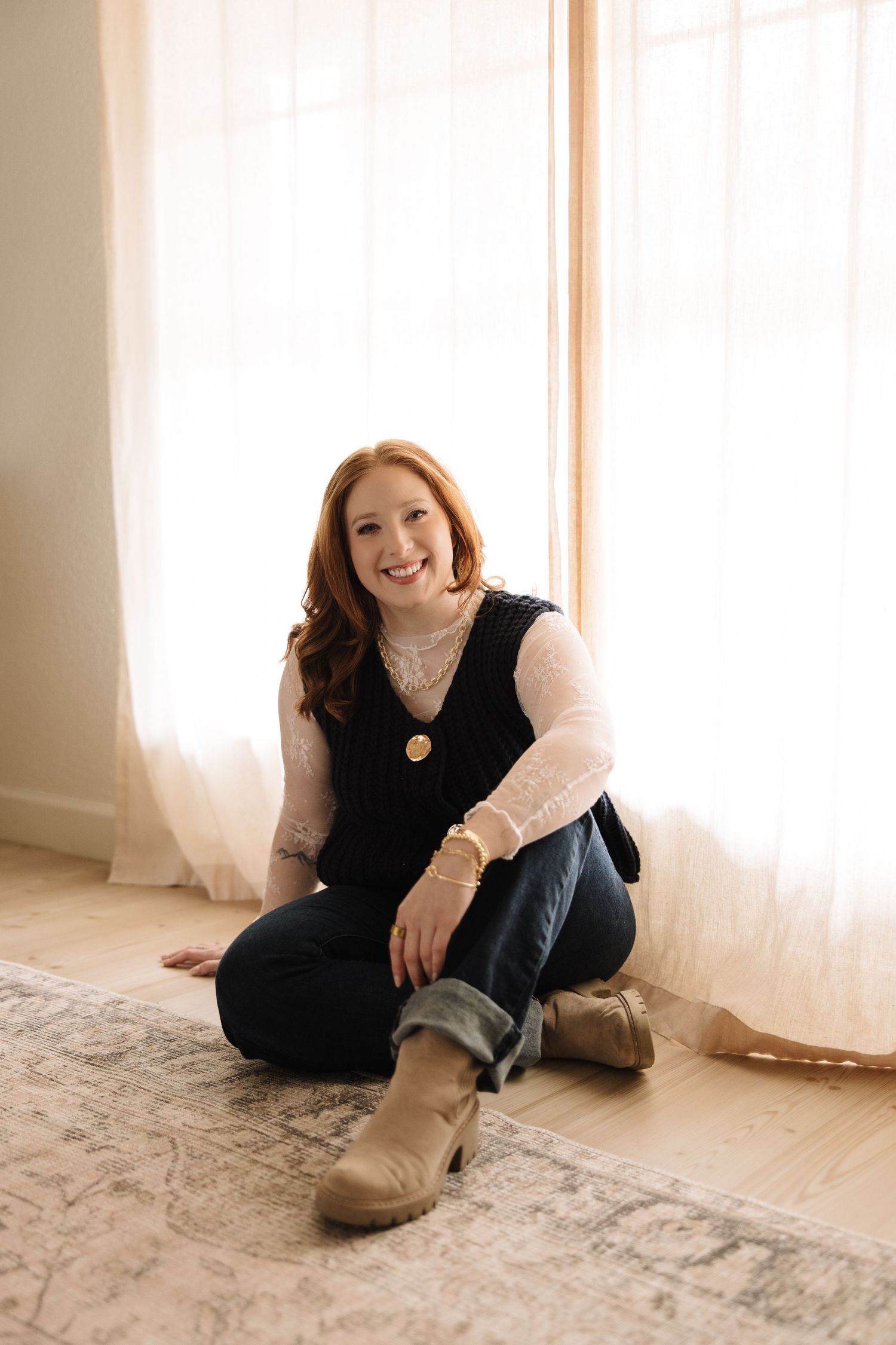 A person in a black sleeveless top and jeans sits casually on a light-colored carpet near sunlit windows.