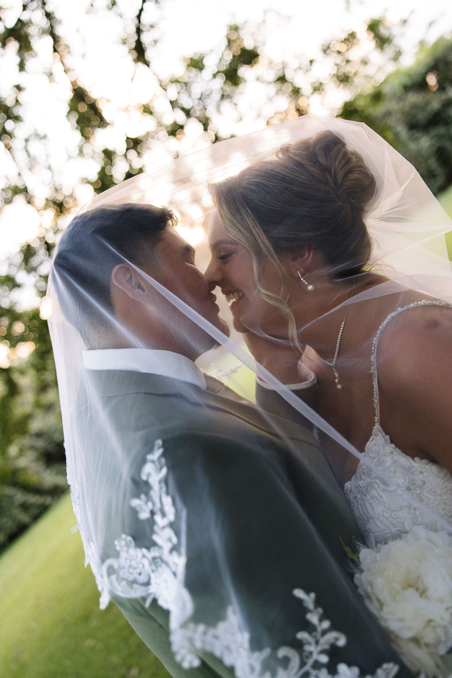 Bride and groom share a romantic moment under a lace-trimmed veil, smiling at each other outdoors on their wedding day.