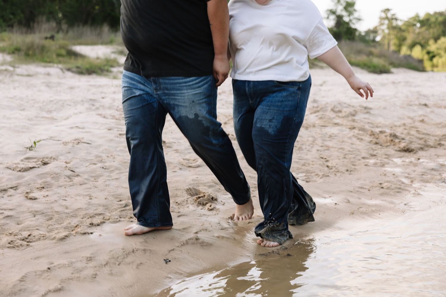 Two people walking barefoot hand in hand through shallow water on a sandy beach wearing jeans.