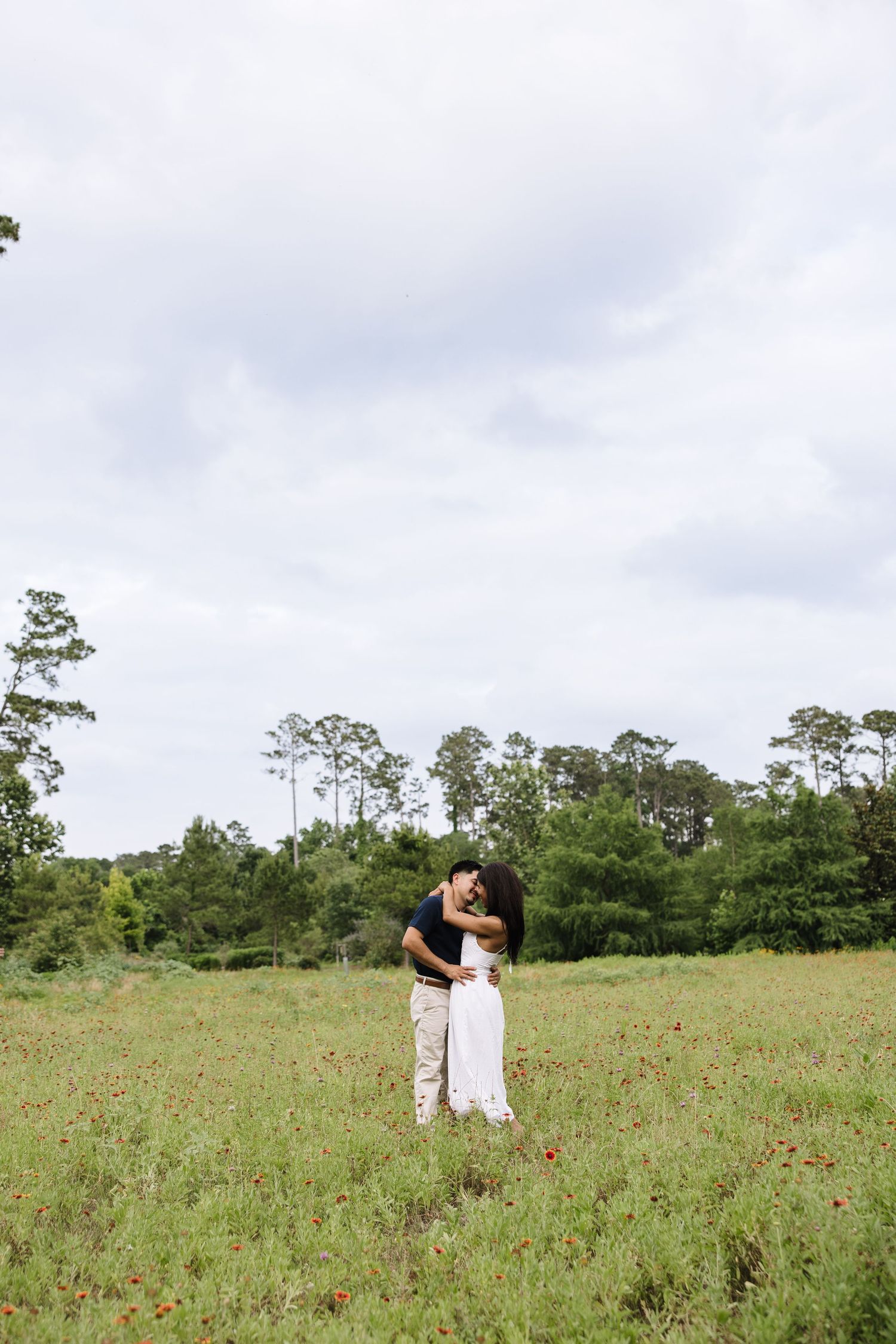 Couple embracing in a wildflower meadow with tall trees and cloudy sky in the background.