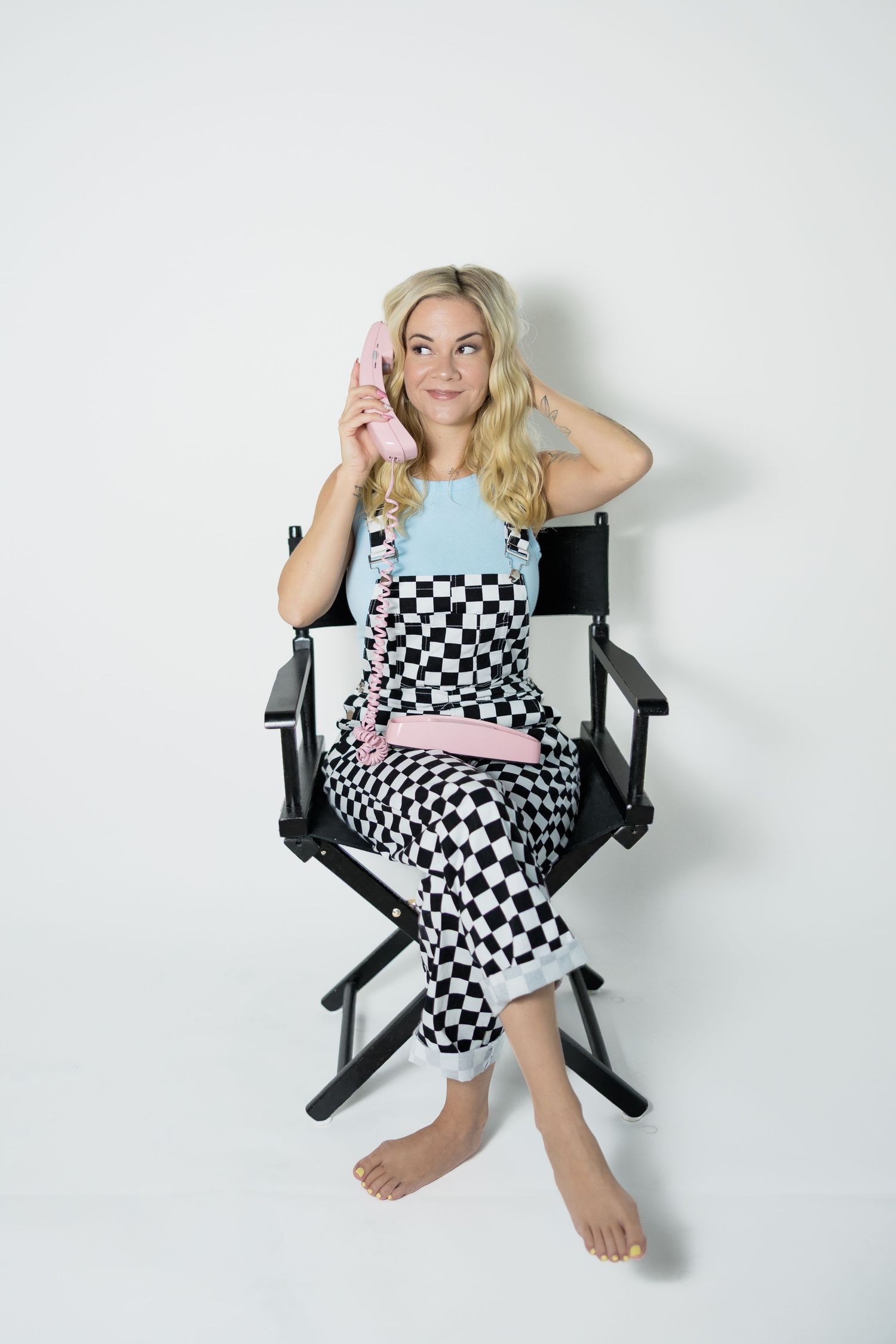 Person seated in director's chair wearing checkered jumpsuit against white backdrop.