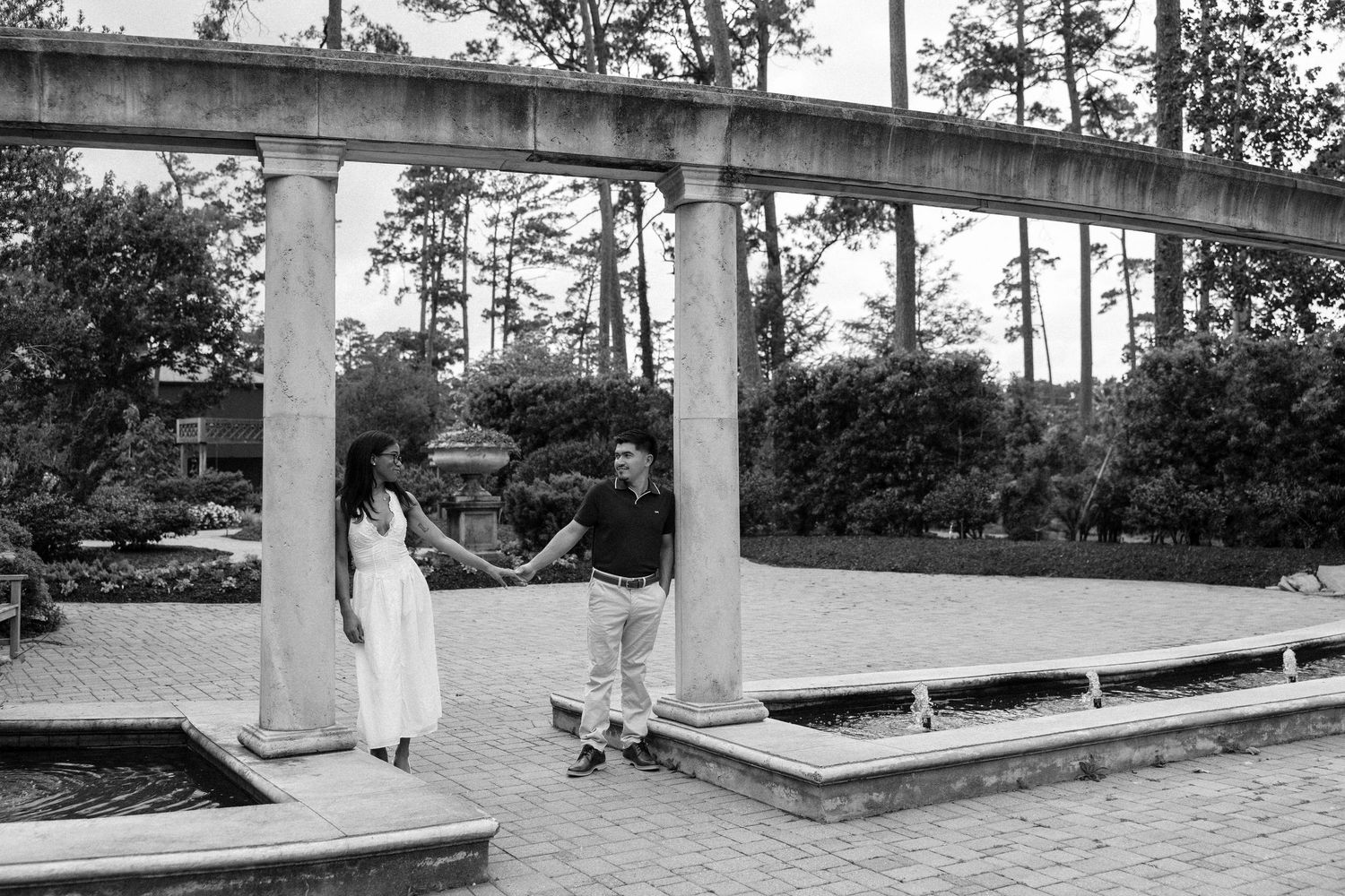 Black and white photo of a couple holding hands near elegant stone columns by a reflective pond.