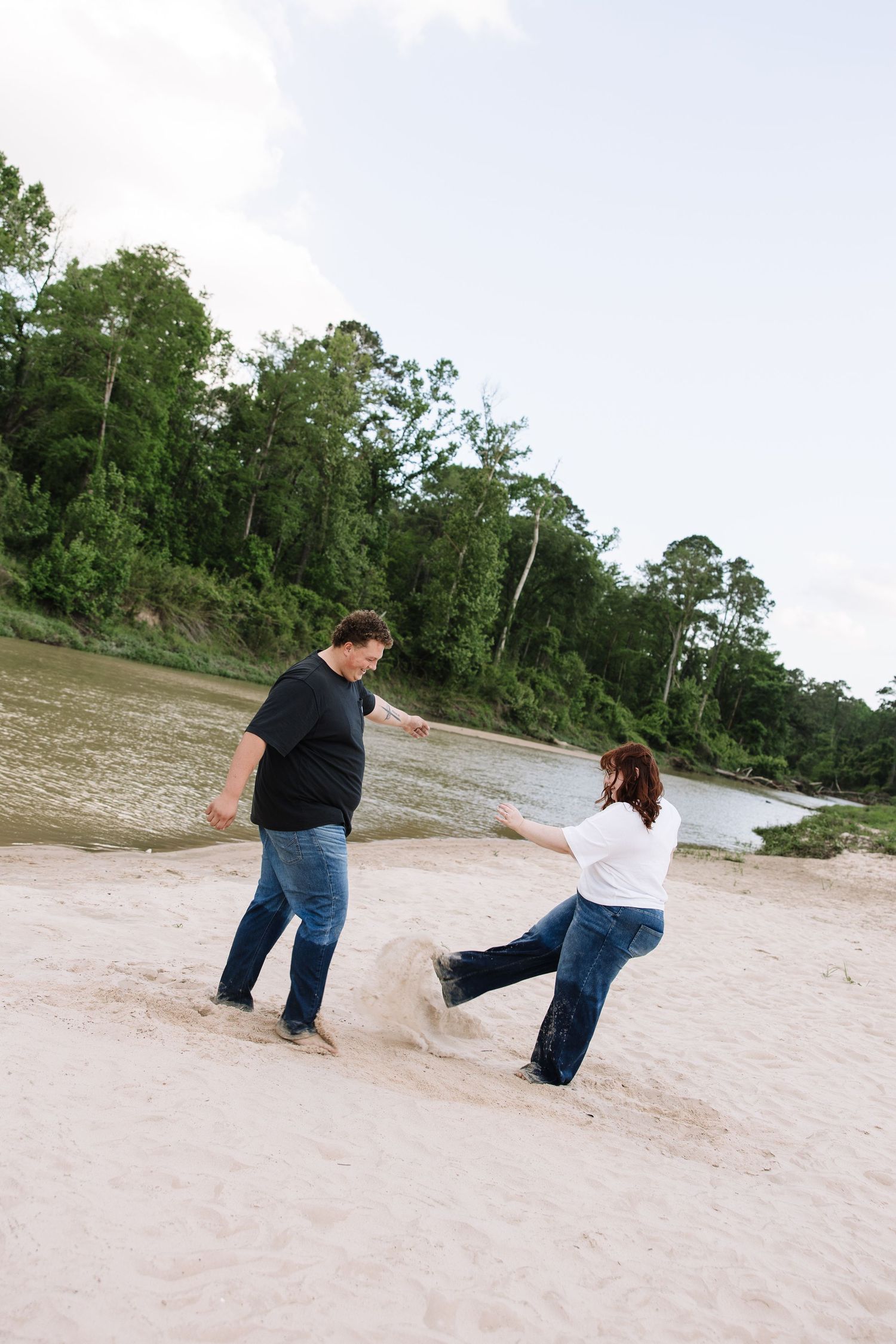 Couple playfully dancing on sandy beach with lush green trees and calm river in the background.
