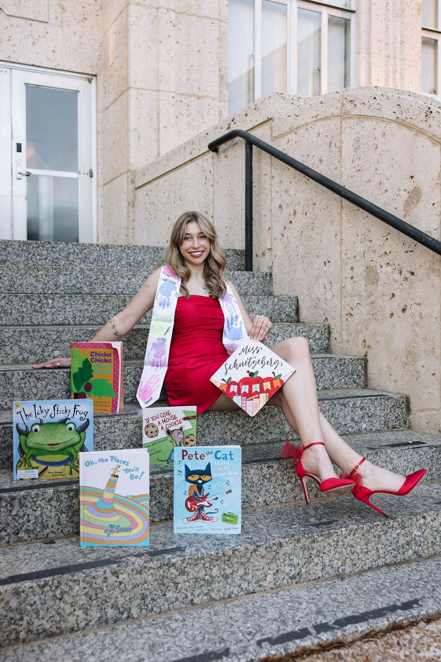 Smiling graduate in red dress and heels sits on stone steps surrounded by colorful children's books including Pete the Cat.