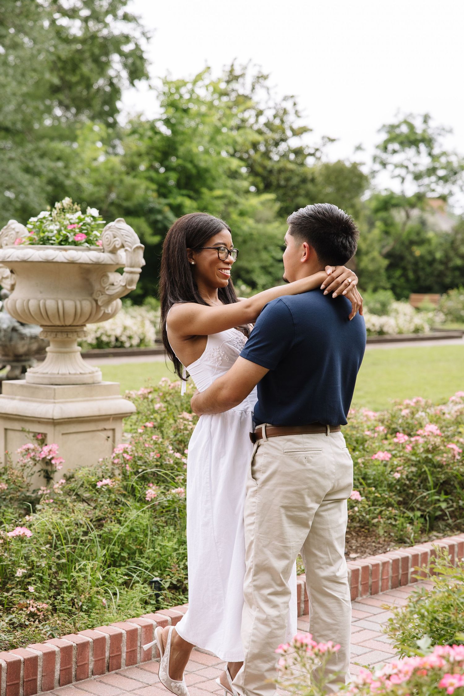Happy couple embracing and smiling at each other in a beautiful rose garden with a decorative stone urn nearby.