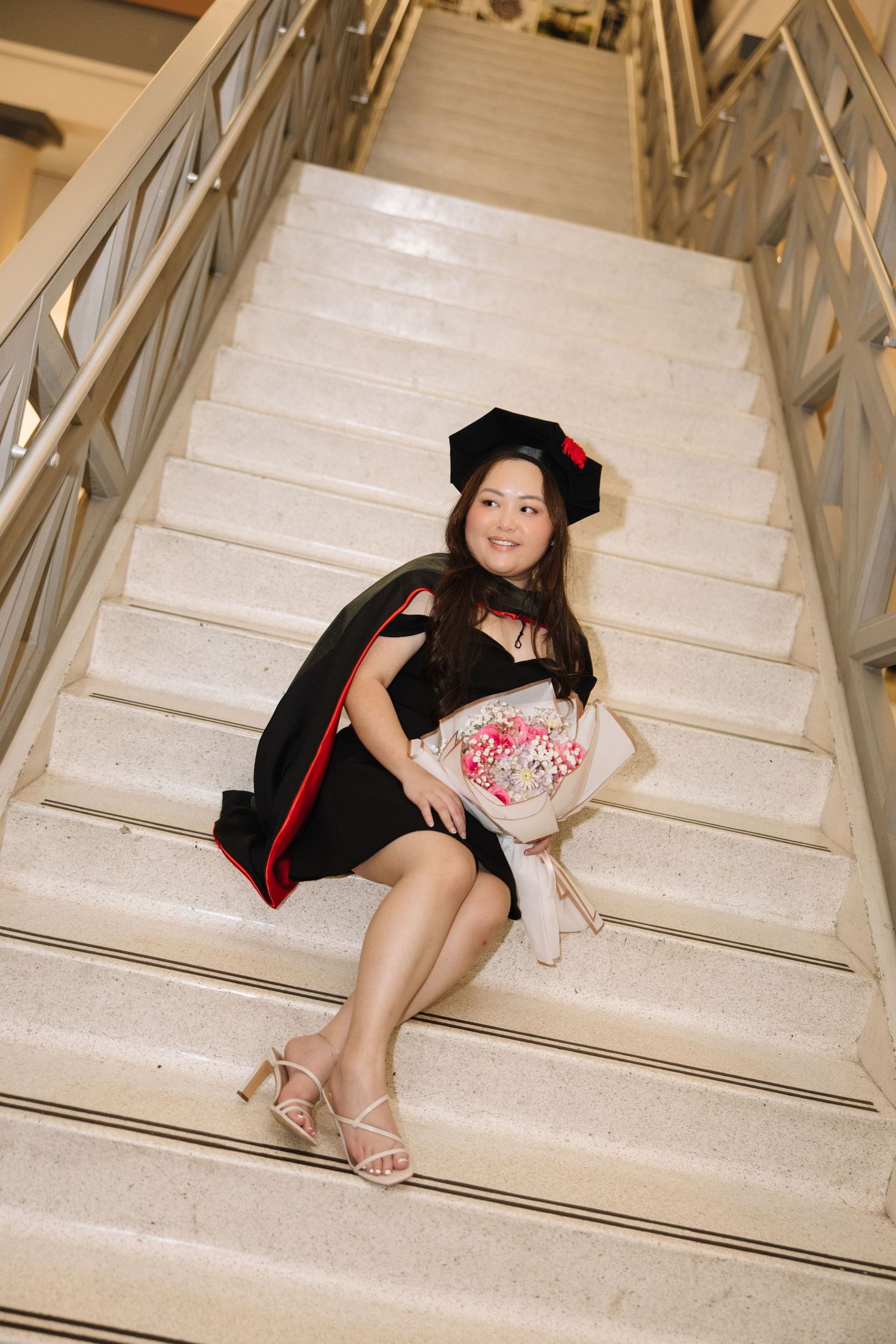 Young woman in black graduation gown and cap sits on marble stairs holding a floral bouquet, celebrating her achievement.