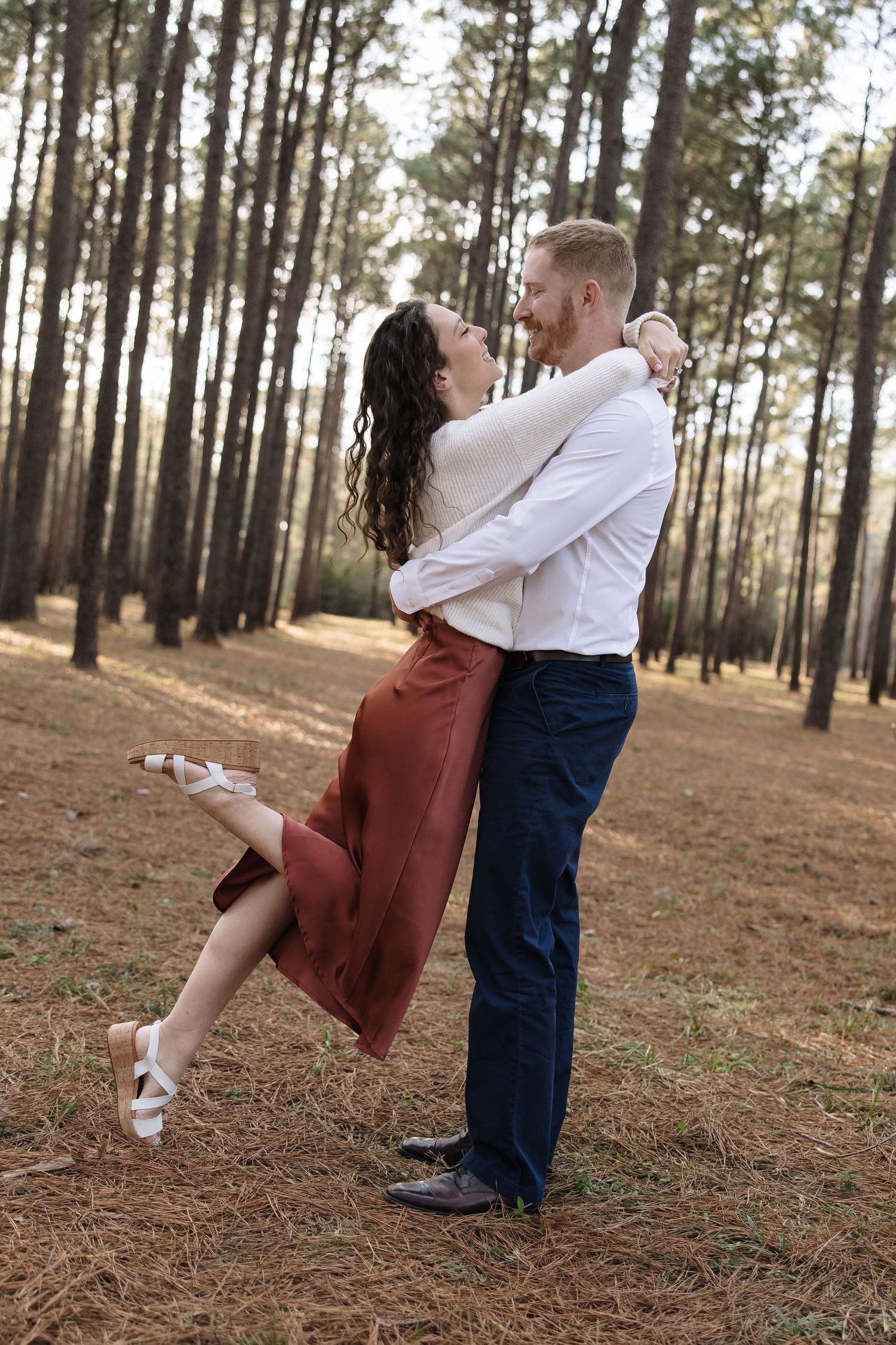 A couple dances playfully on a pine needle covered forest slope at sunset.