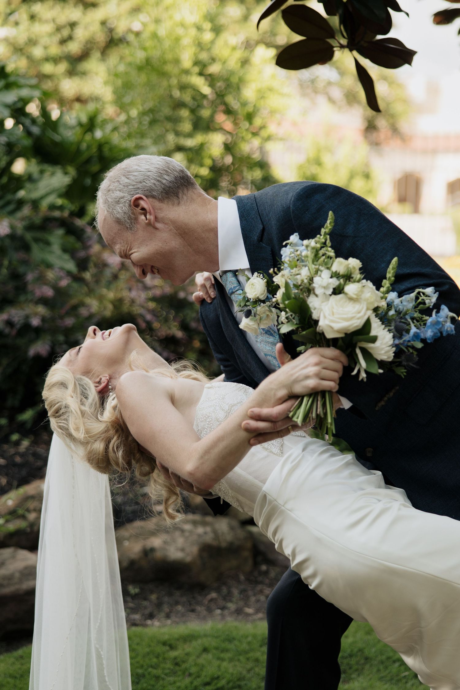 Couple in wedding attire performs romantic dip pose in garden setting.
