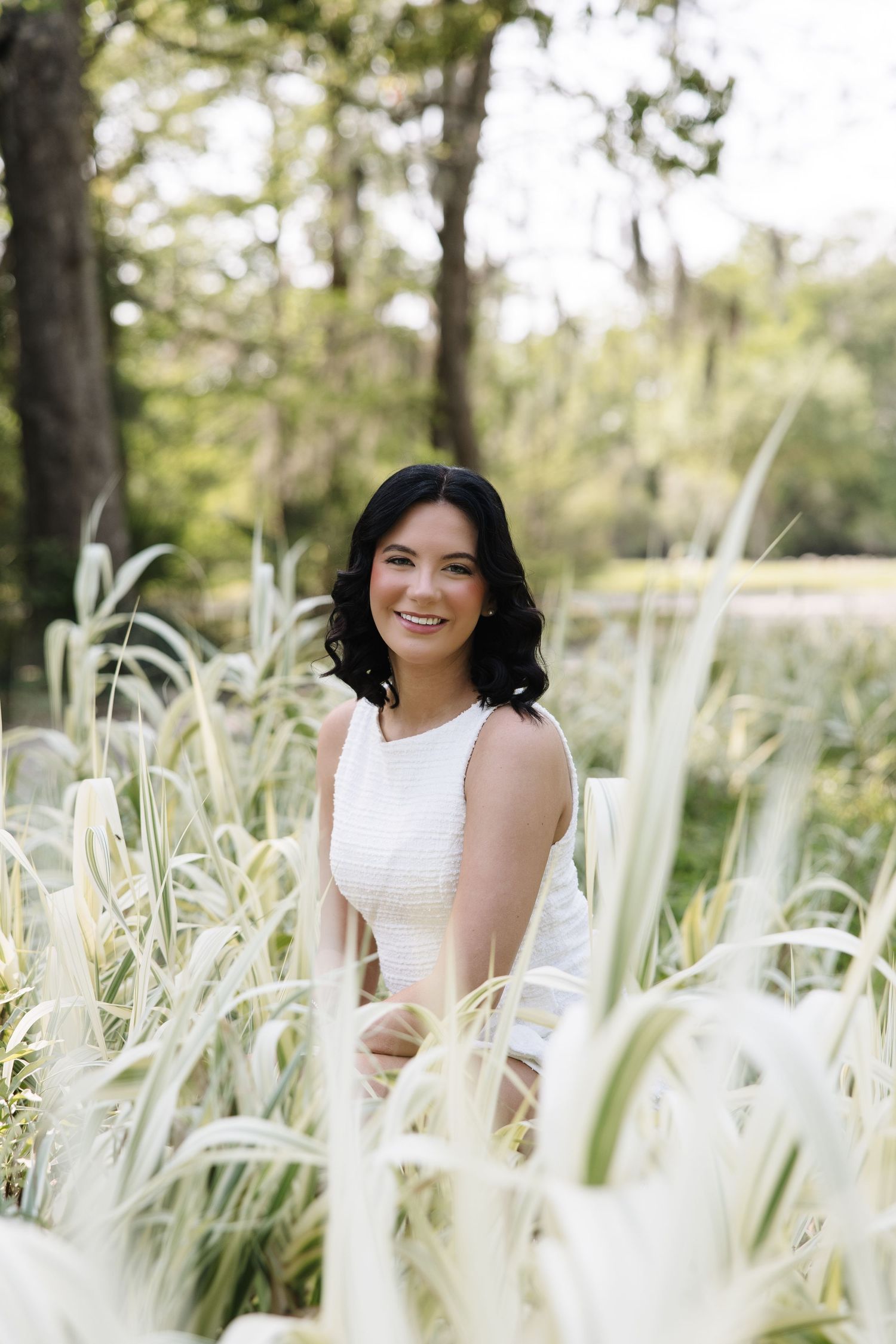 Woman in white dress smiling among tall ornamental grasses in a lush green outdoor park setting.