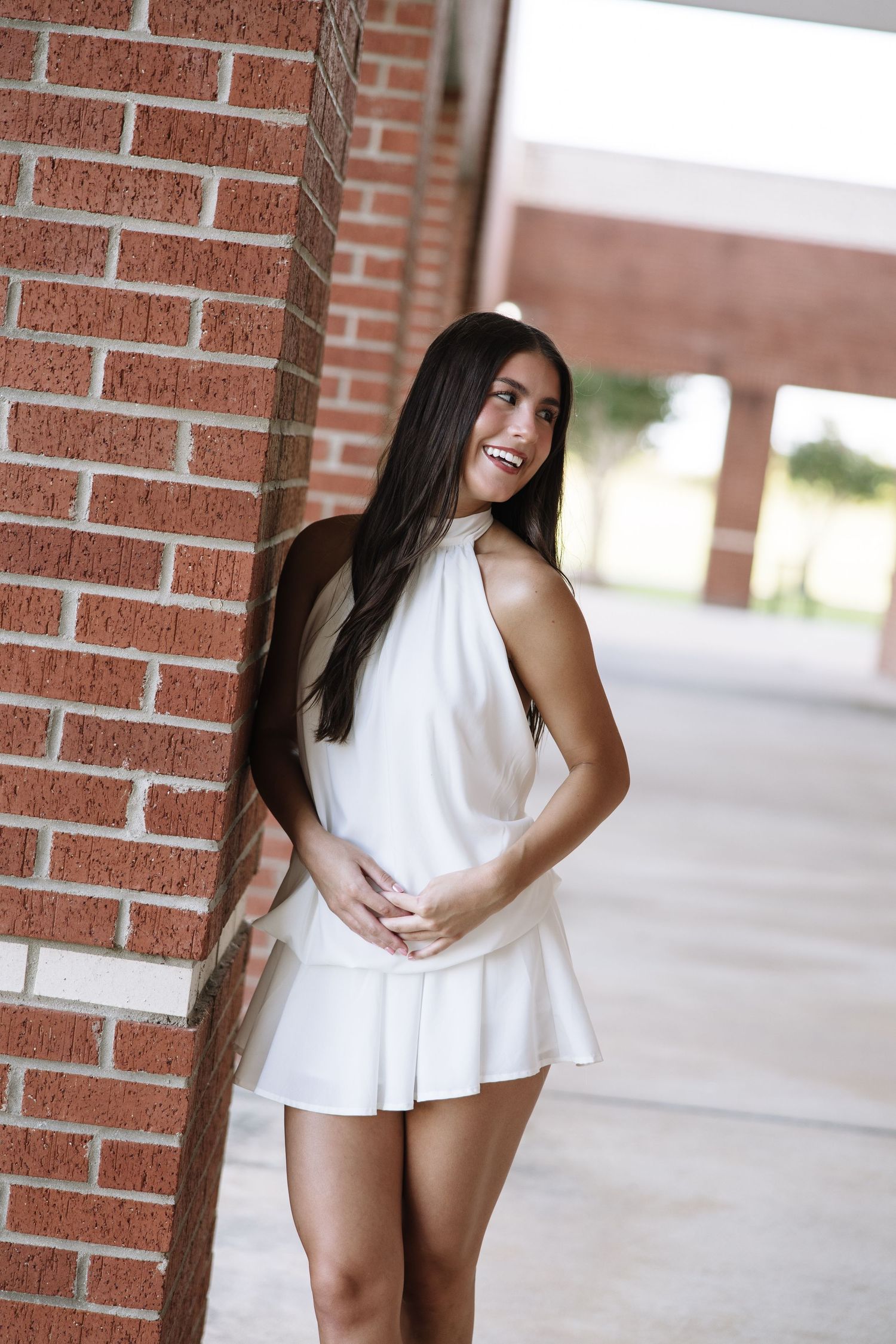 Young woman in white halter dress smiling and leaning against a red brick wall outdoors.
