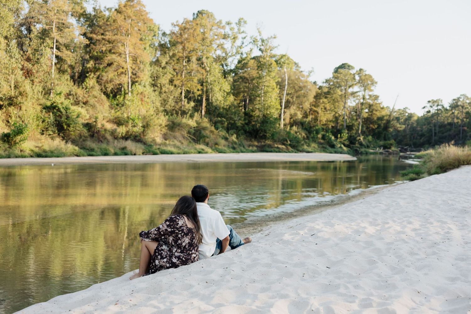 People enjoying a scenic autumn day along a tranquil river bank surrounded by tall pine trees and golden foliage.