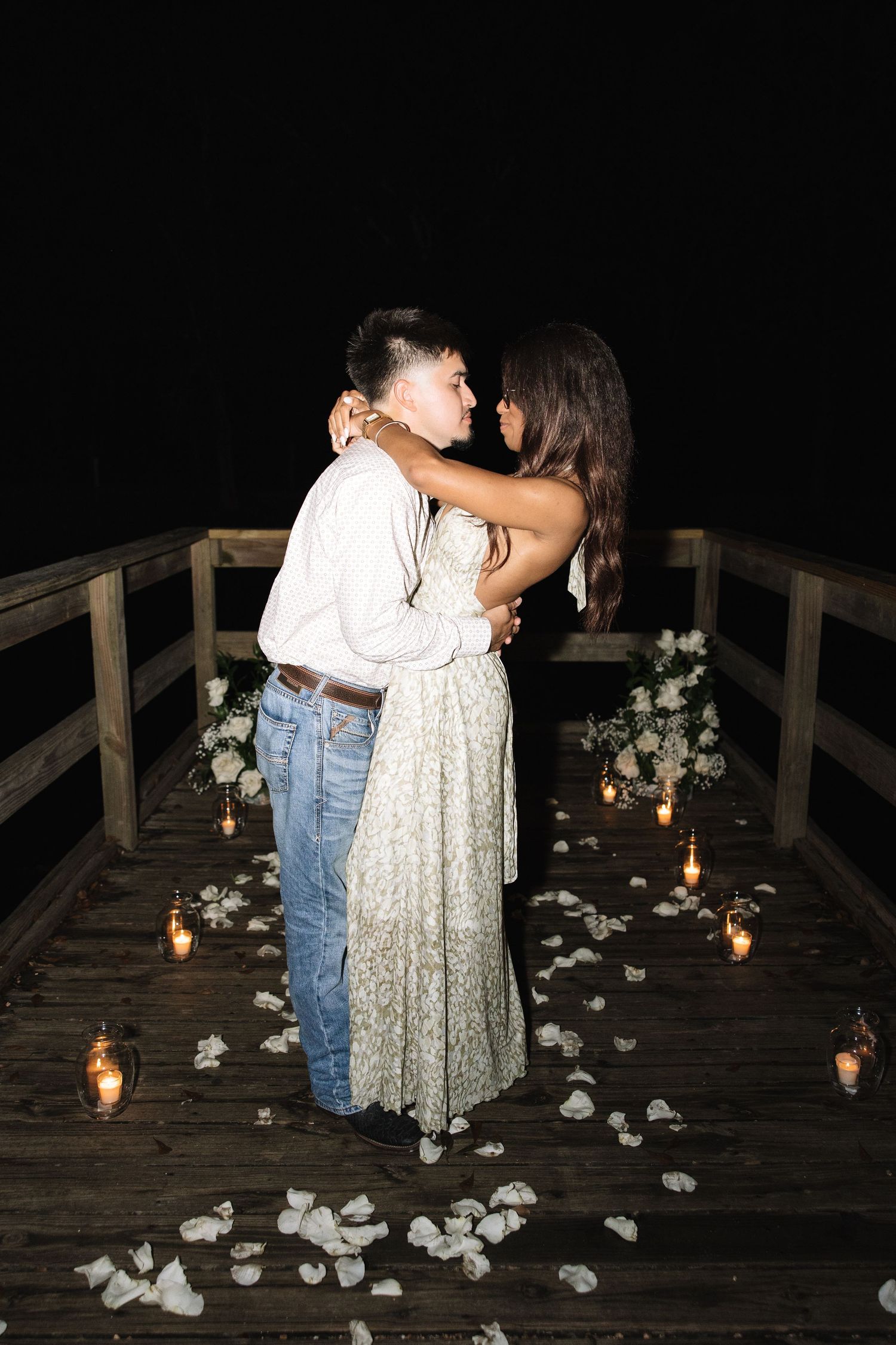 A romantic proposal scene on a wooden deck lit by candlelight with rose petals scattered on the ground.