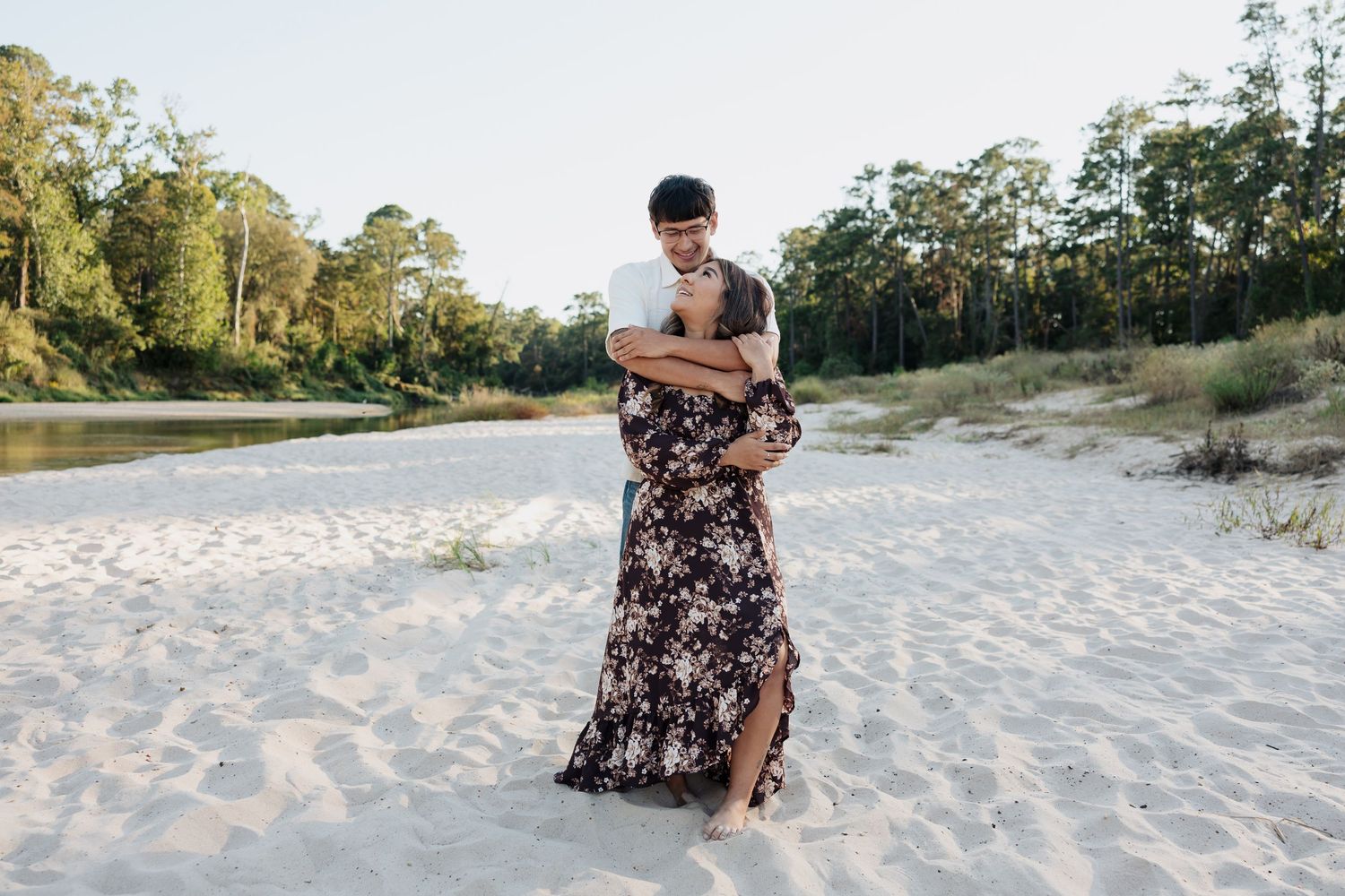A person in a floral dress walks along a sandy beach path through coastal vegetation at sunset.
