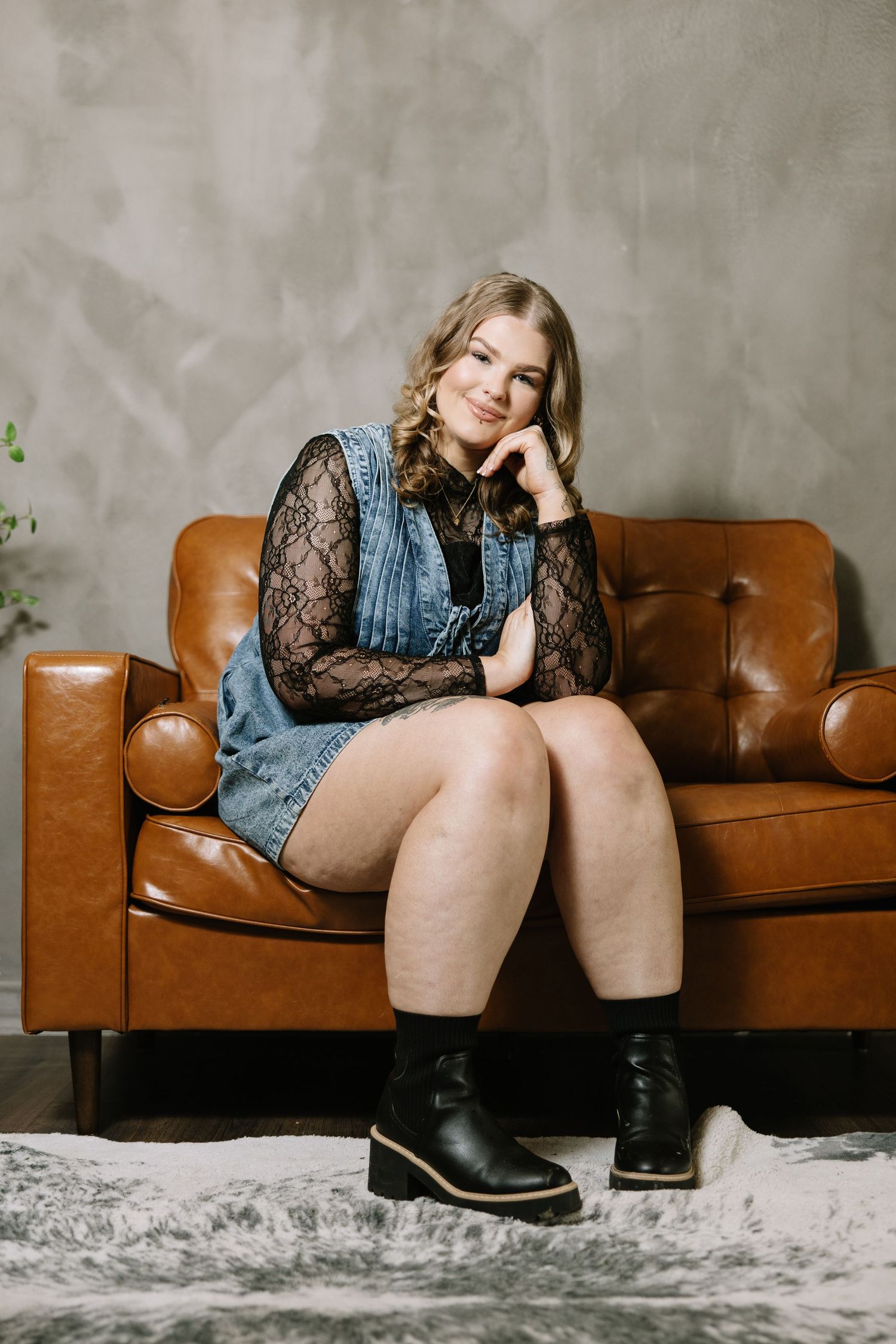 Person in denim dress and lace sleeves sitting on brown leather couch against textured wall.