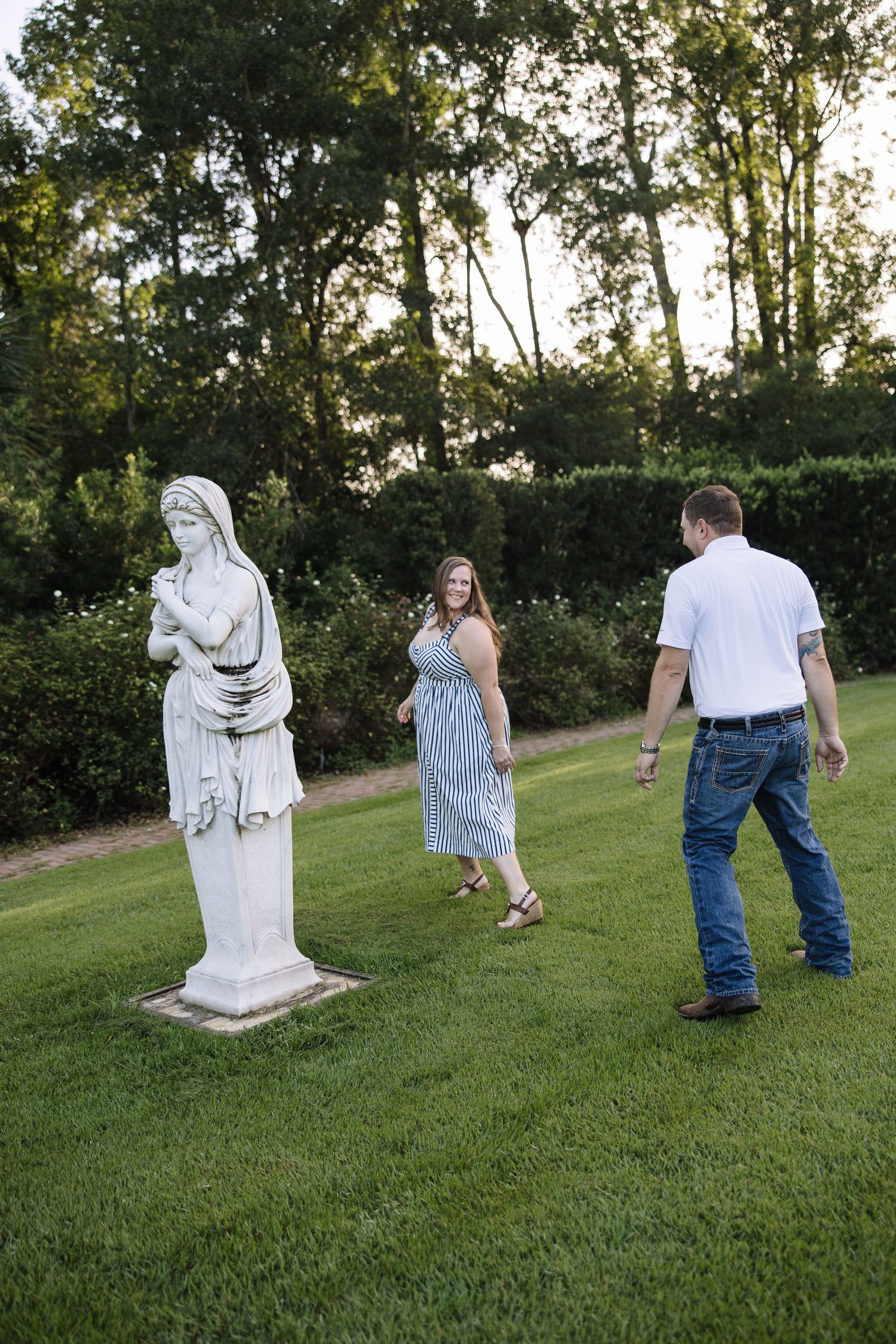 Couple walking near a classical white statue in a lush green garden at dusk.
