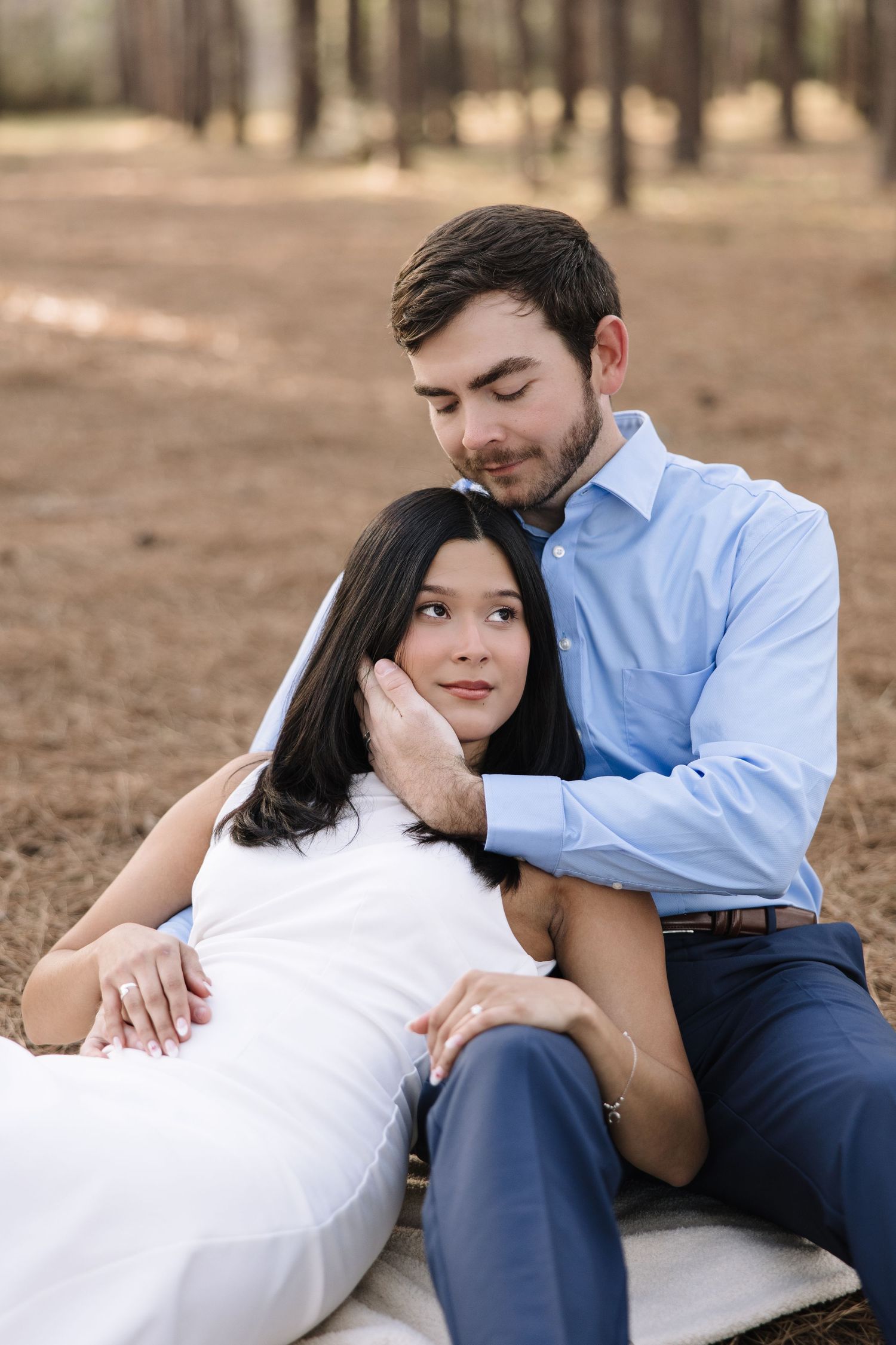 Romantic couple in white and blue outfits share an intimate moment while resting on blanket in a wooded setting.
